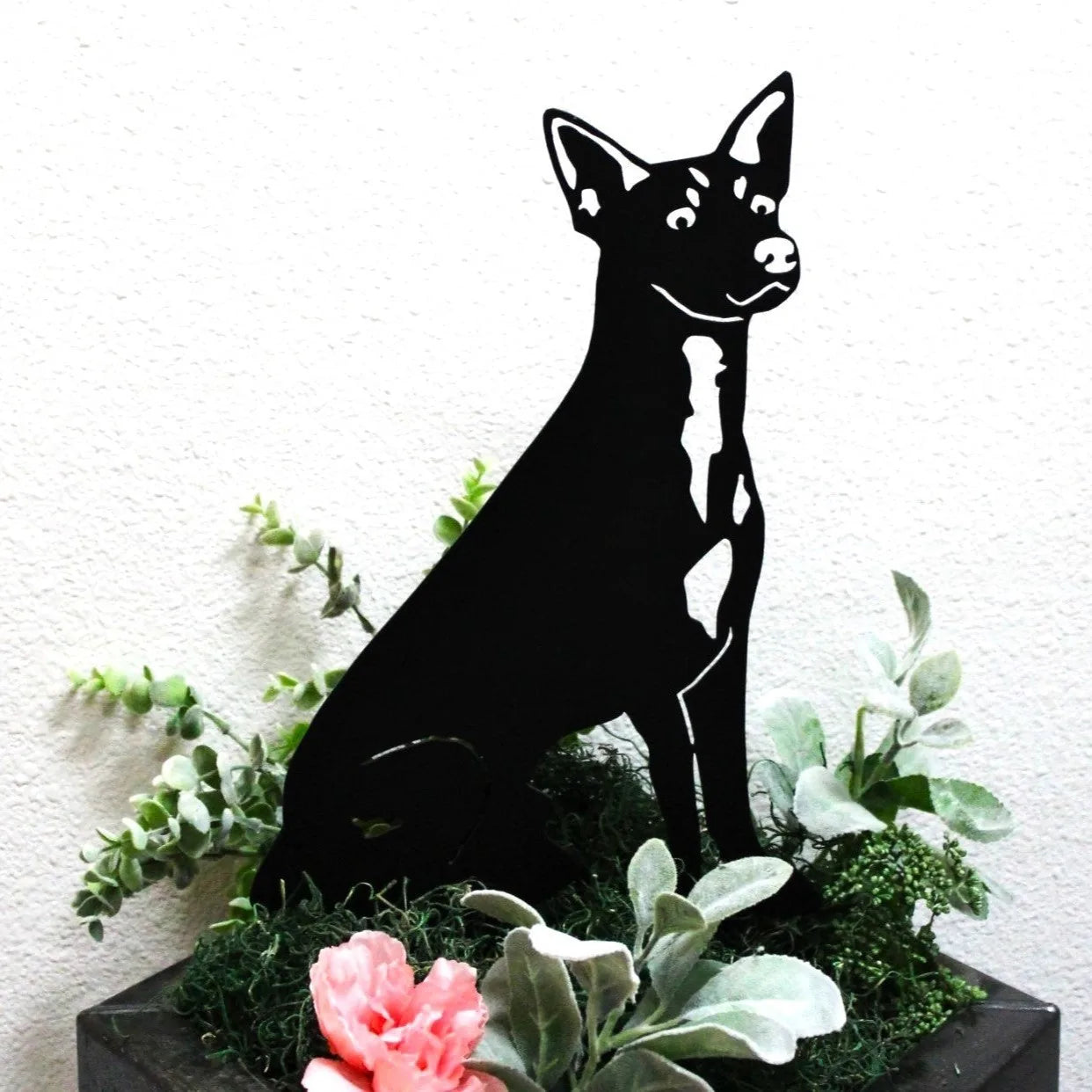 Black metal silhouette of a Rat Terrier seated among greenery and pink flowers in a decorative planter.
