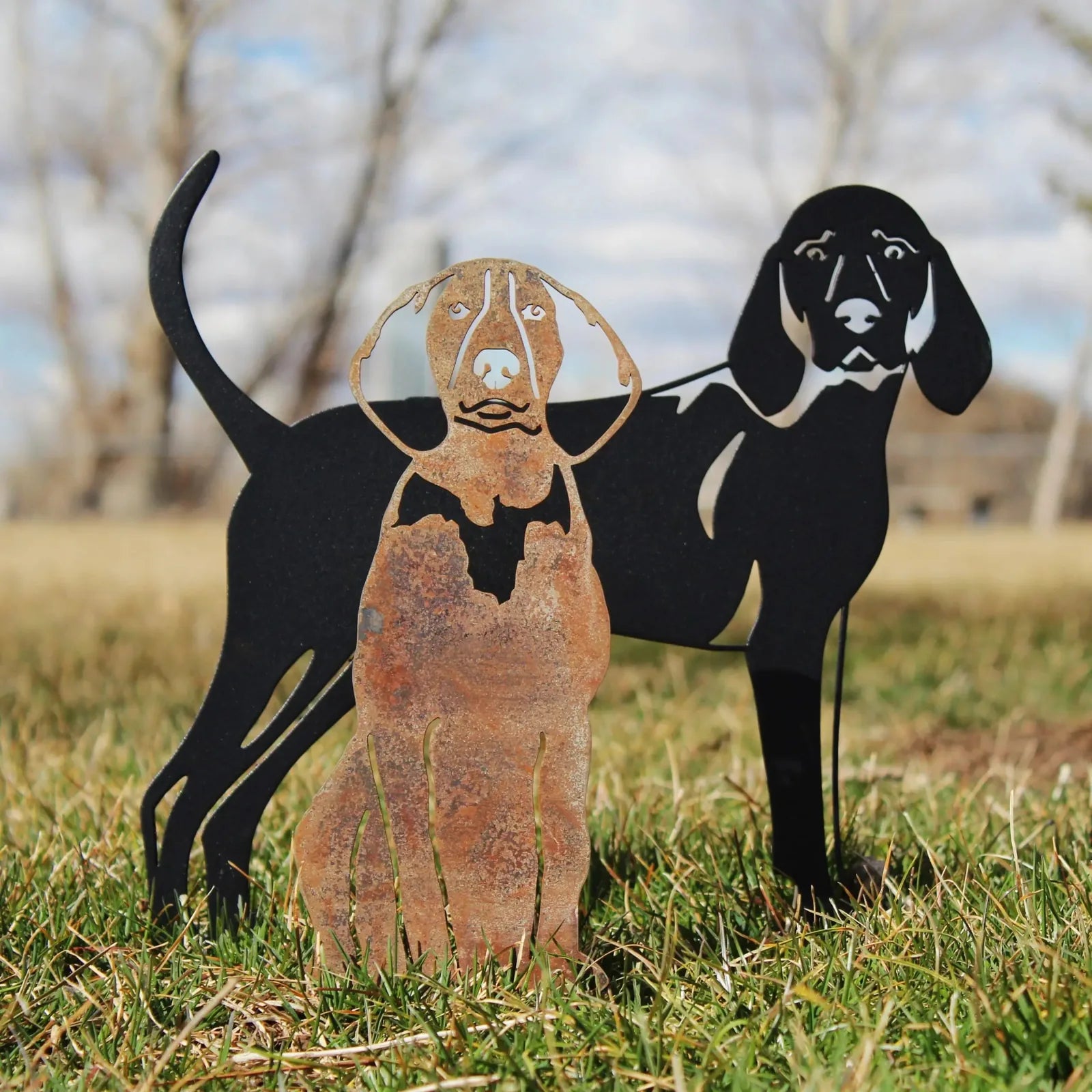 Metal silhouette of two Redbone Coonhounds, one rusted Corten steel, one matte black, standing on grass.