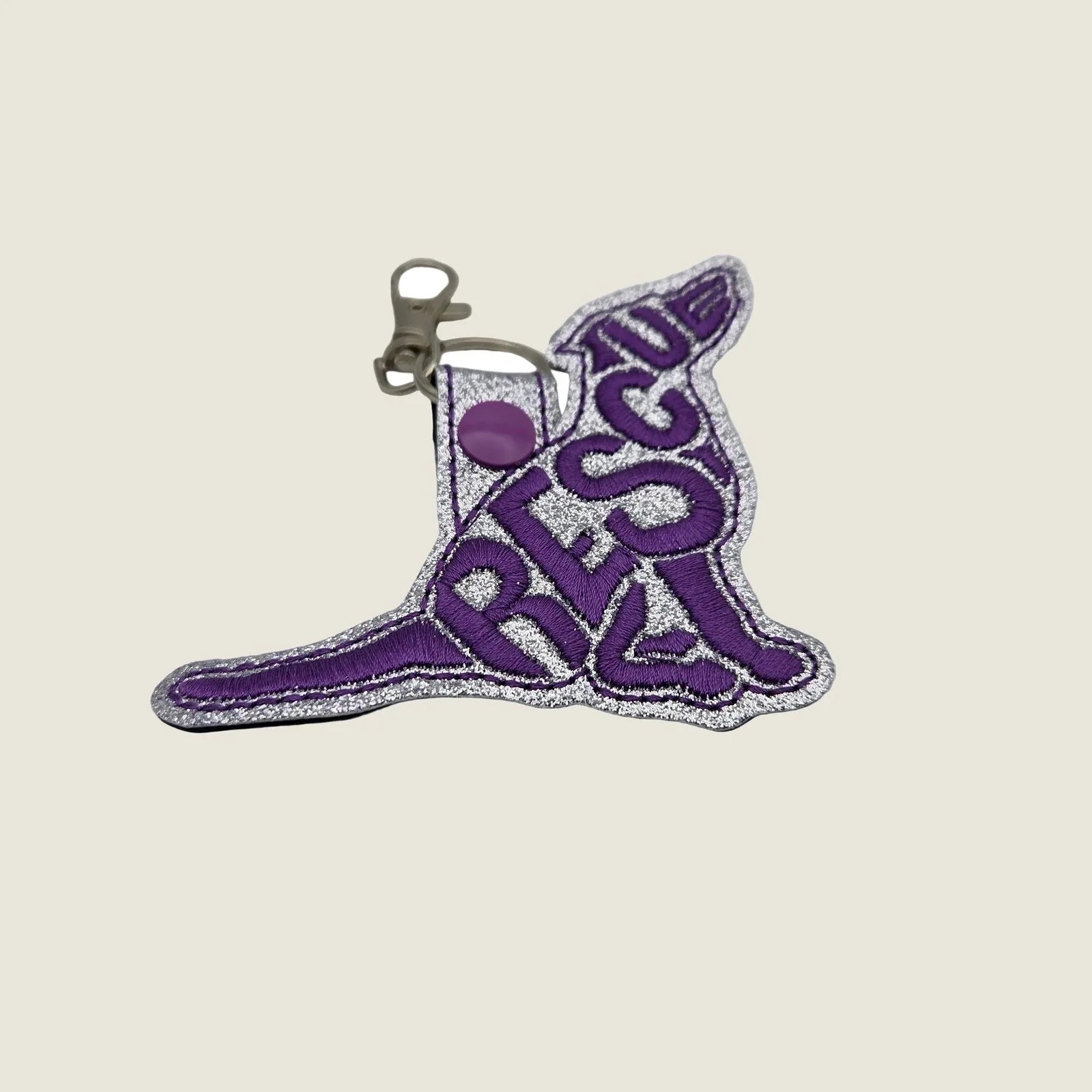 Embroidered purple "RESCUE" keychain tab with silver glitter backing and lobster clasp for attaching to bags or keys.