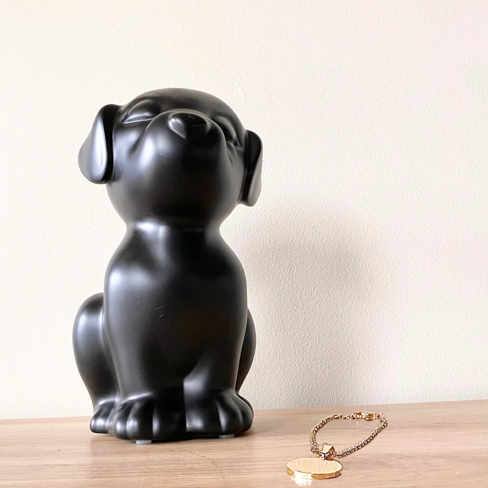 Black retriever dog urn with a smooth, matte finish, featuring a gold pendant nearby on a wooden surface.