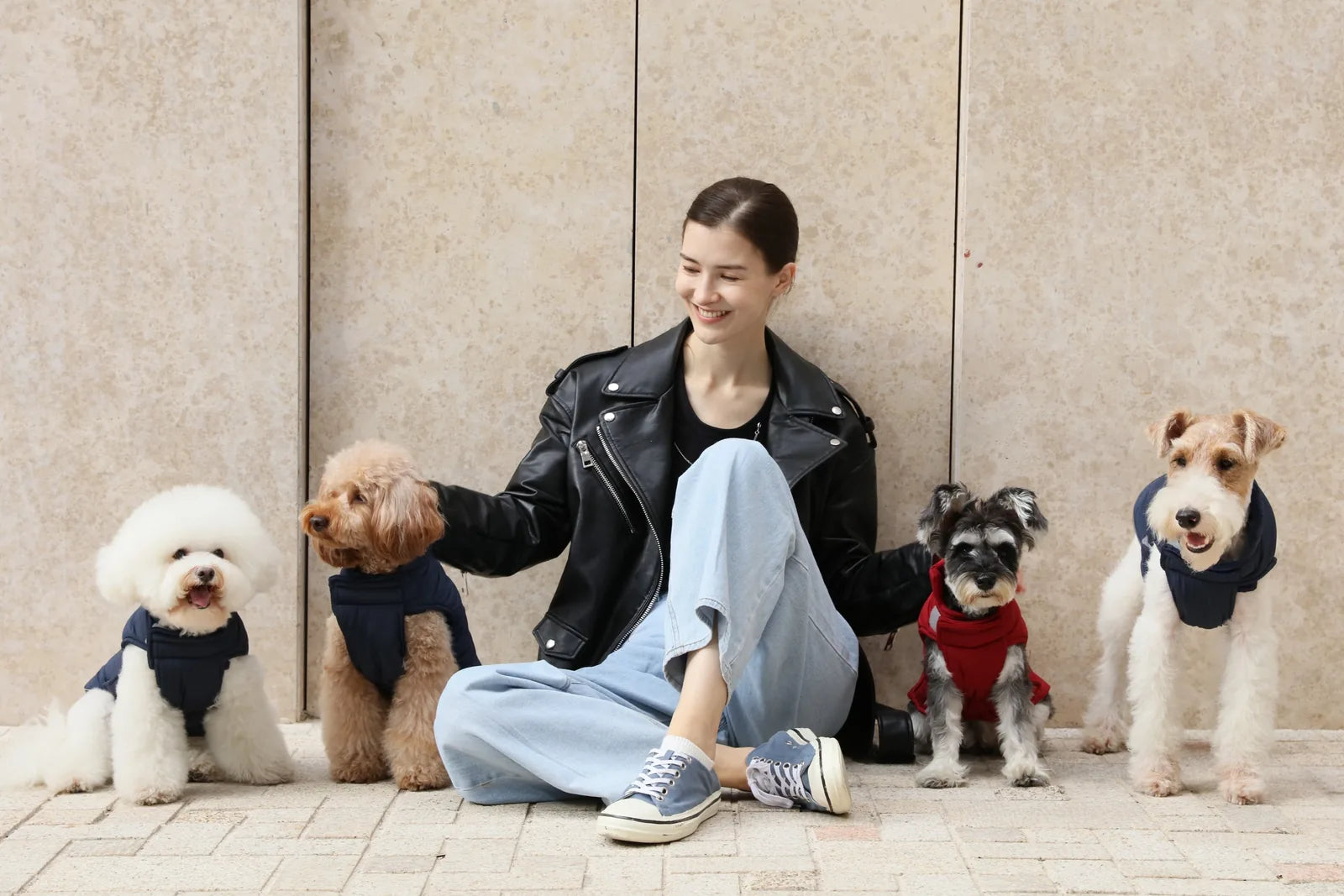 Four dogs in stylish jackets sit beside a smiling woman, showcasing the reversible red dog vest jacket against a neutral b...