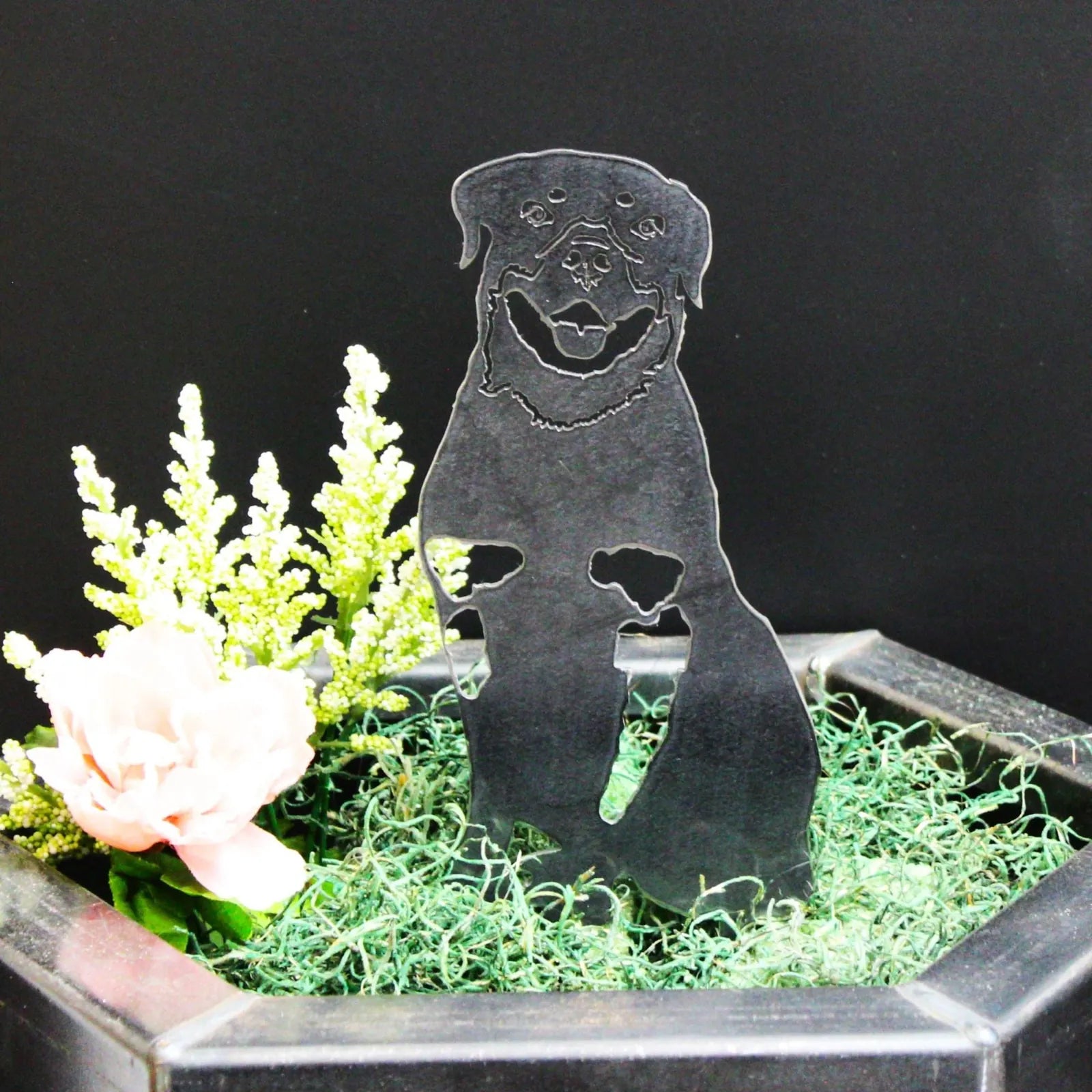 Black metal silhouette of a Rottweiler sitting among green moss and flowers, perfect for outdoor dog decor.