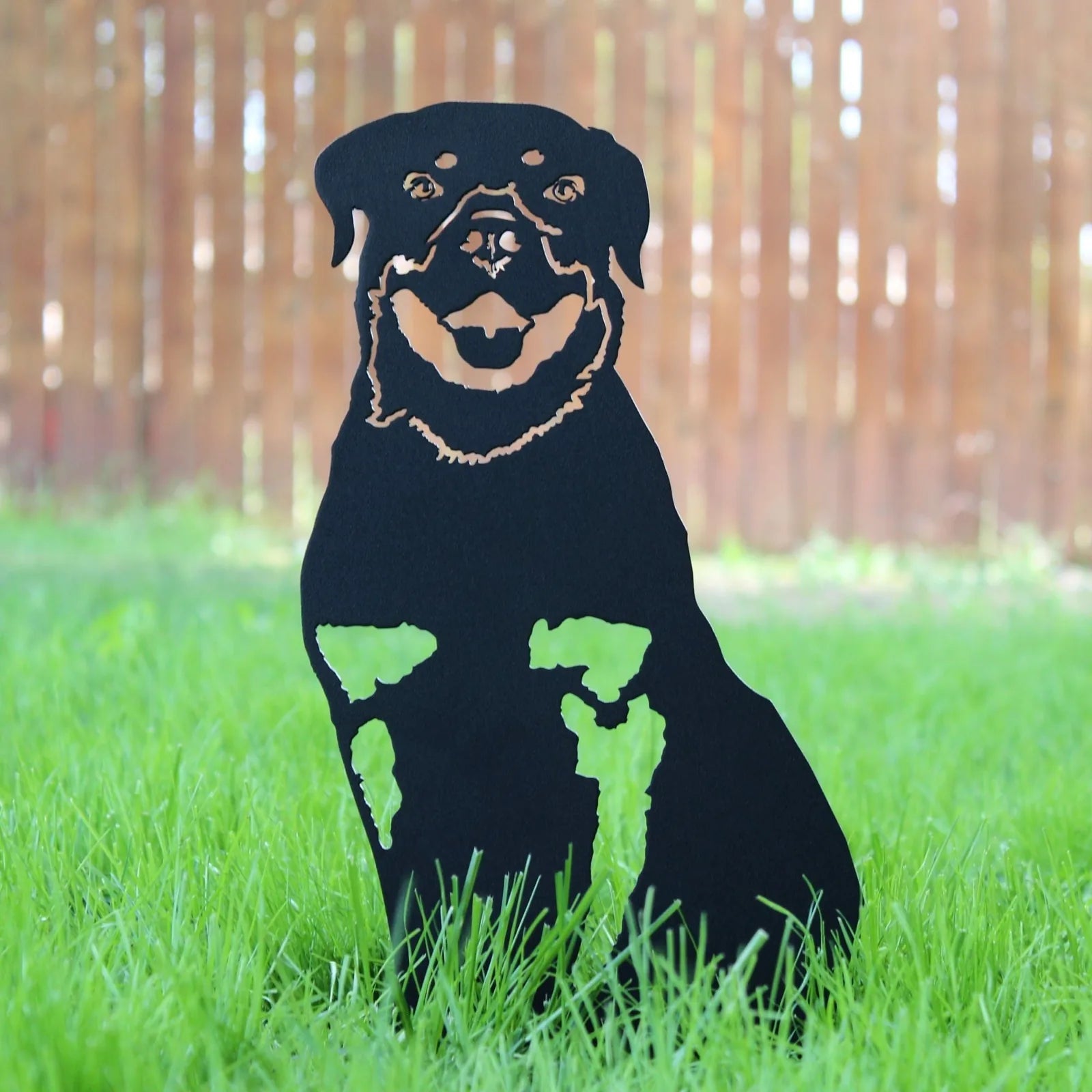 Black metal Rottweiler silhouette decor in grass, featuring a smiling expression against a wooden fence backdrop.