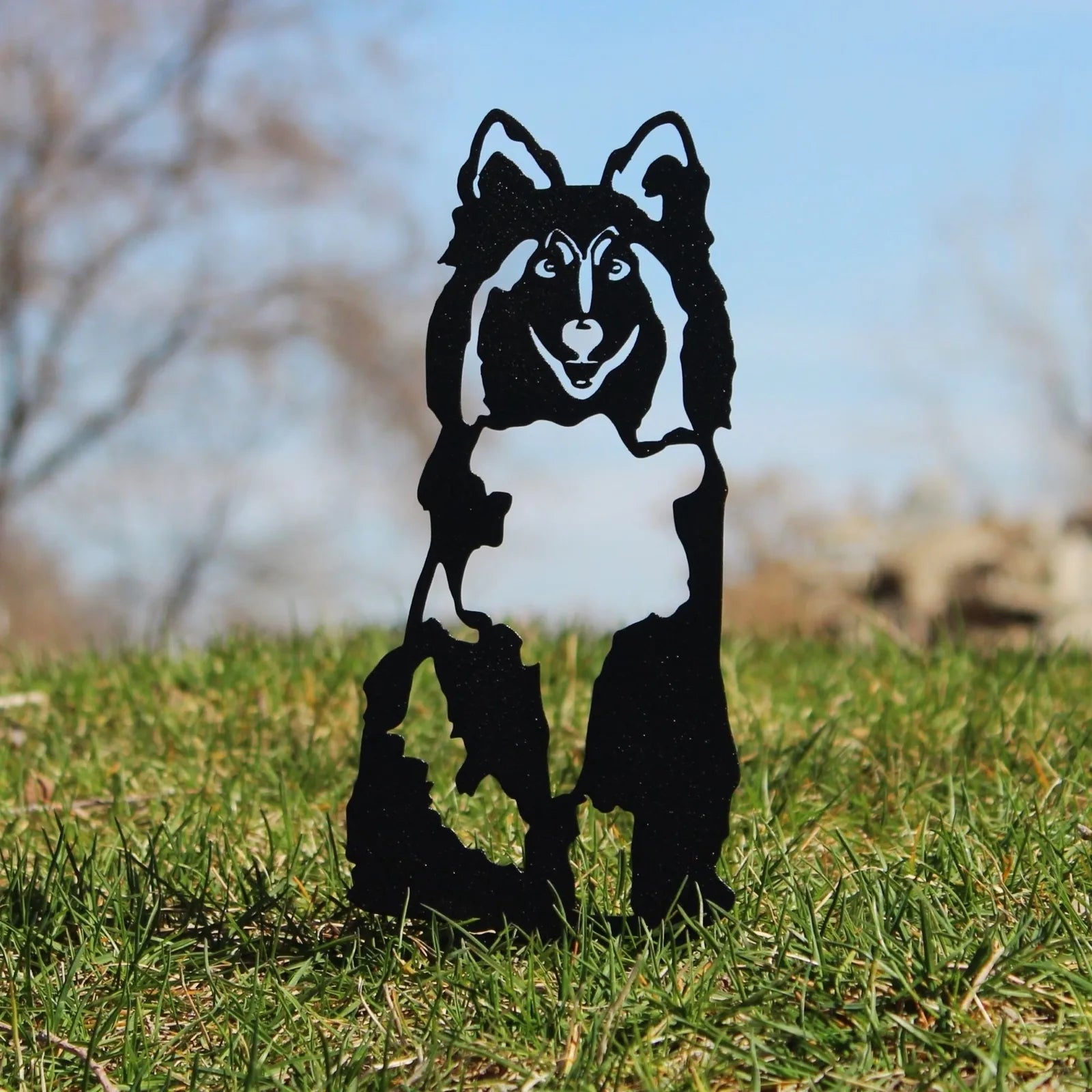 Black metal silhouette of a Rough Collie dog standing on grass, reflecting sunlight against a blue sky background.