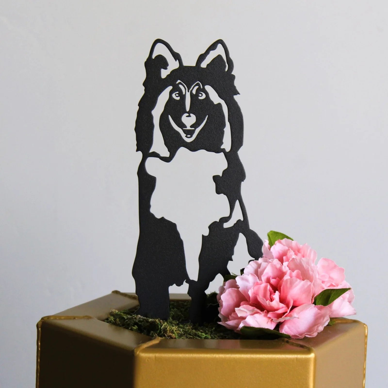 Black Rough Collie metal silhouette sits on a gold pedestal with pink flowers and moss, ideal for outdoor dog decor.