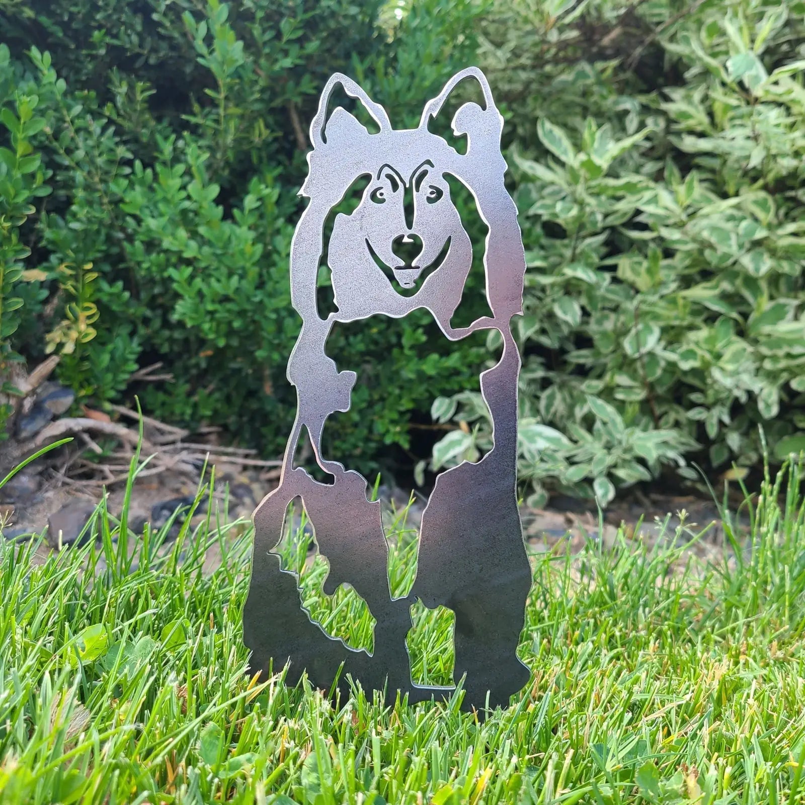 Rough Collie metal silhouette made of black steel, standing in grass with green bushes in the background.