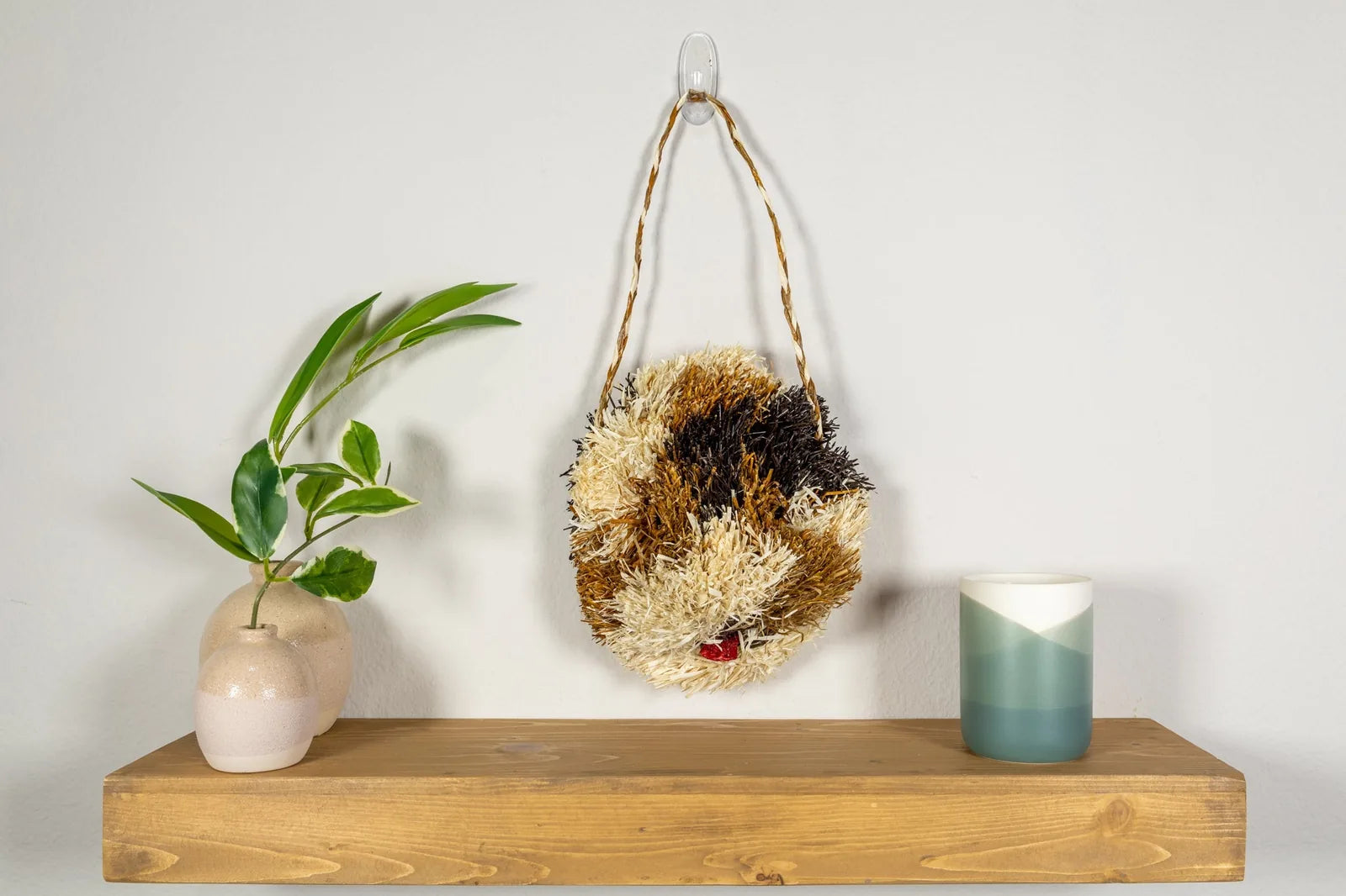 Handwoven Saint Bernard dog mask in natural hues, displayed on a wooden shelf alongside decorative plants and a candle.