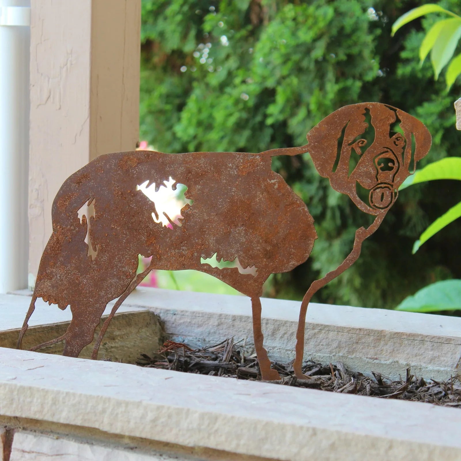 Rusty Corten steel silhouette of a Saint Bernard dog, standing in a garden planter with greenery in the background.