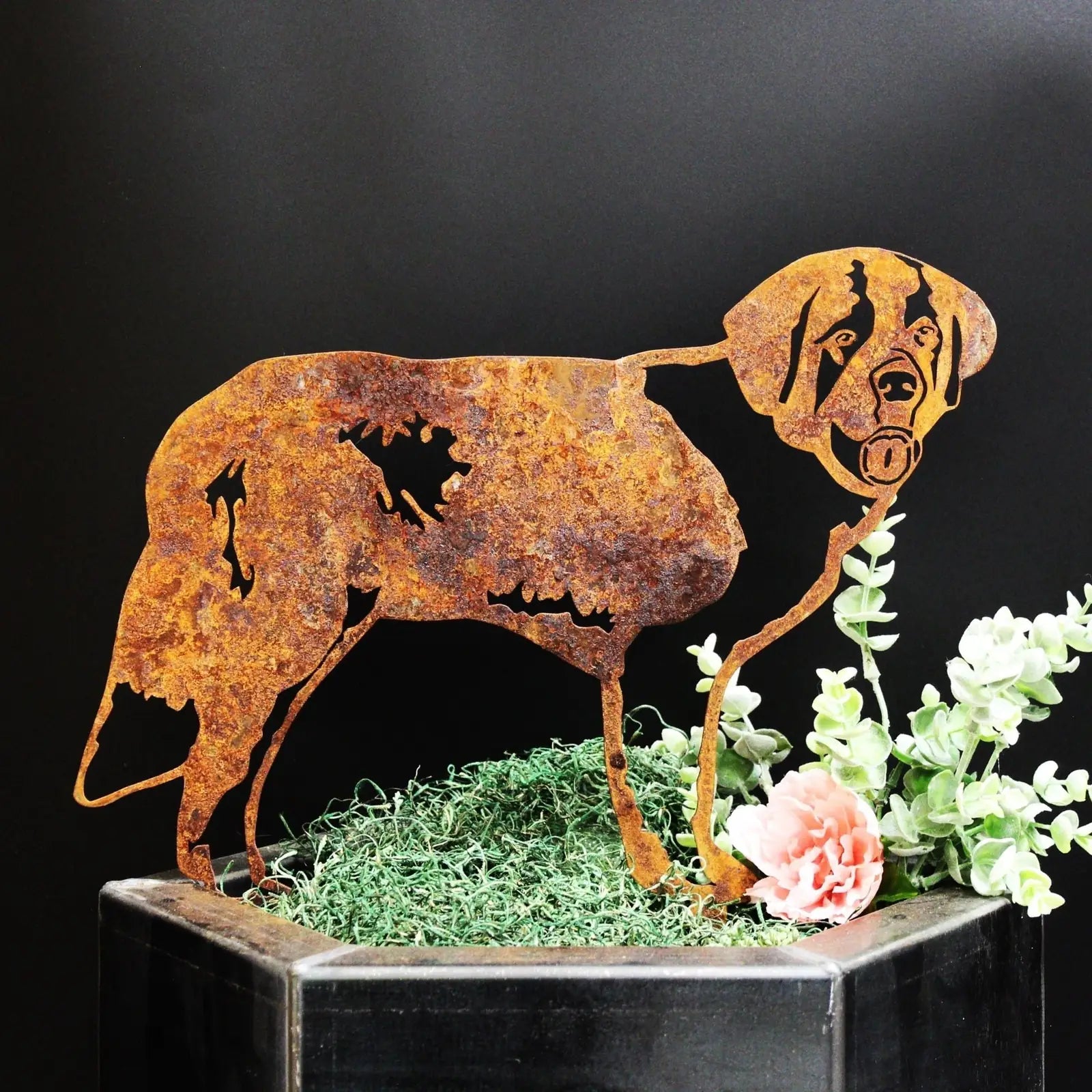 Rust-colored Saint Bernard silhouette made from Corten steel, surrounded by greenery and a pink flower, mounted in outdoor...