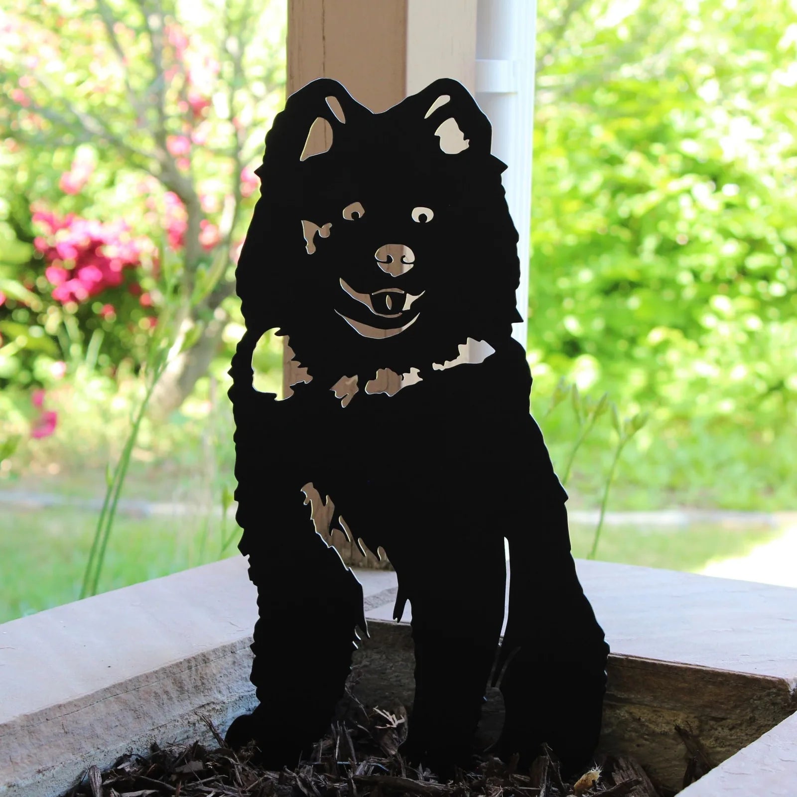 Black Samoyed metal silhouette showcasing playful expression, fitted among garden mulch, with vibrant green backdrop.