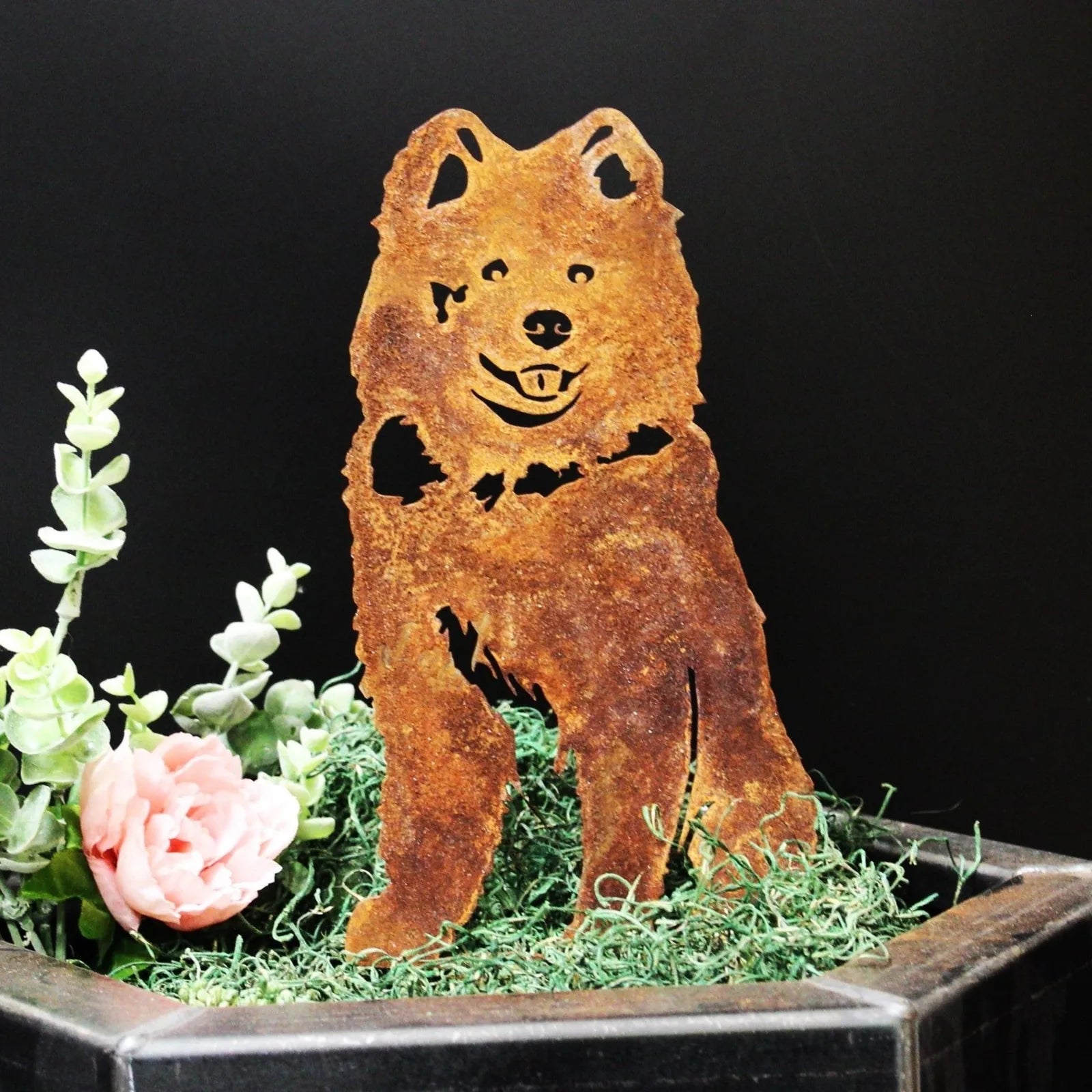 Rustic Samoyed metal silhouette, crafted from Corten steel, displayed among green foliage and pink flowers.