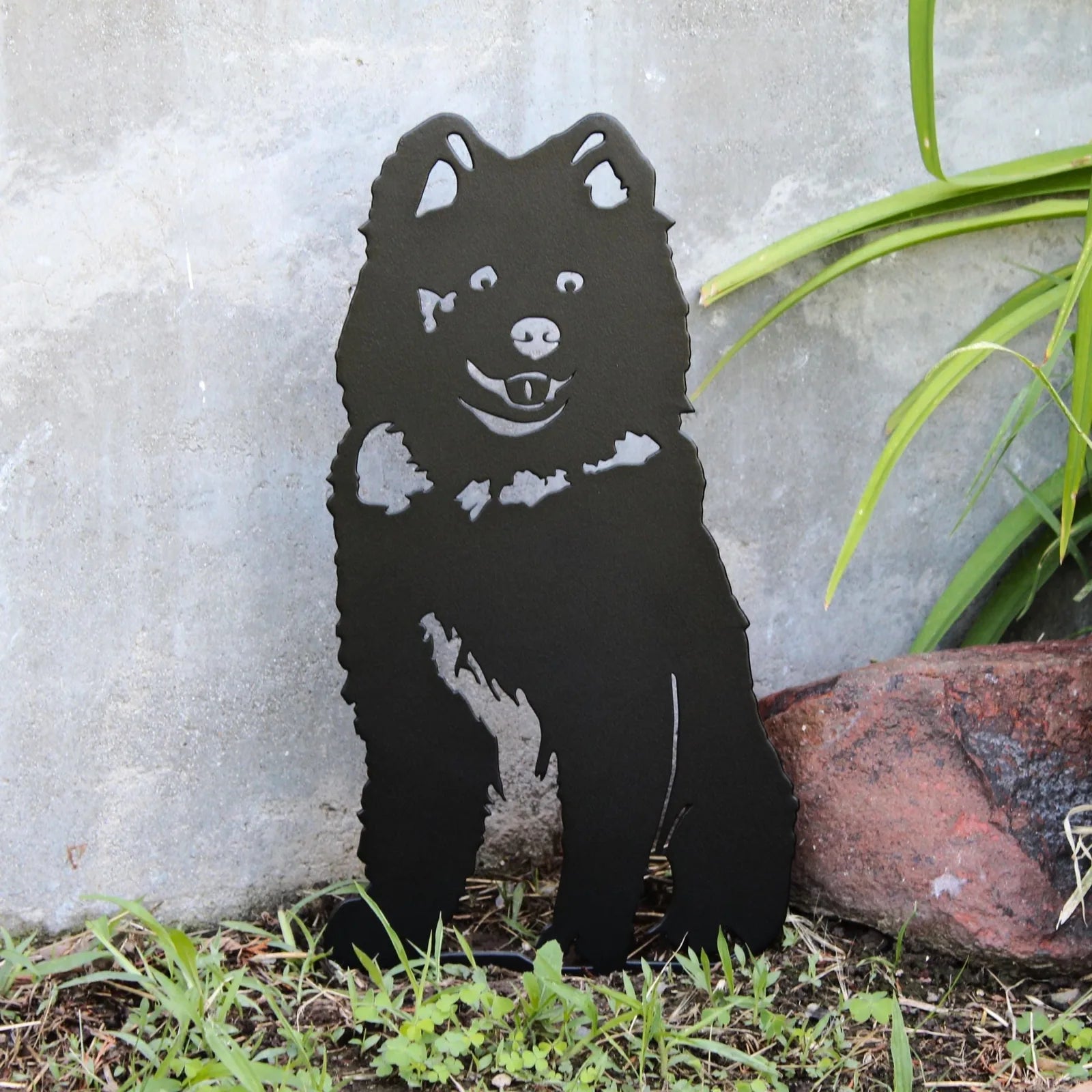 Black metal silhouette of a Samoyed dog, showcasing detailed features, propped in grass near a stone wall.