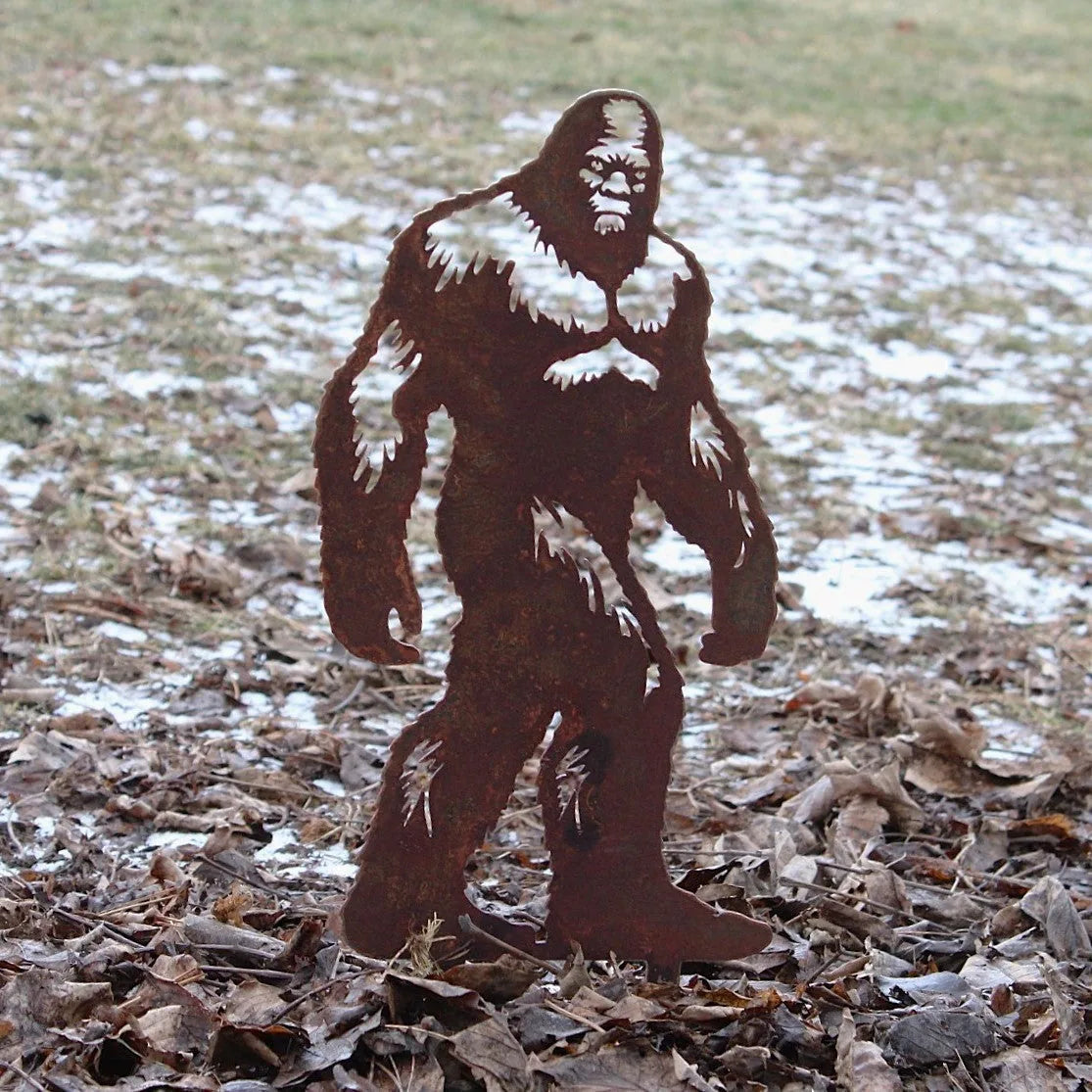 Sasquatch metal silhouette in rusty Corten steel, standing among dried leaves in an outdoor setting.