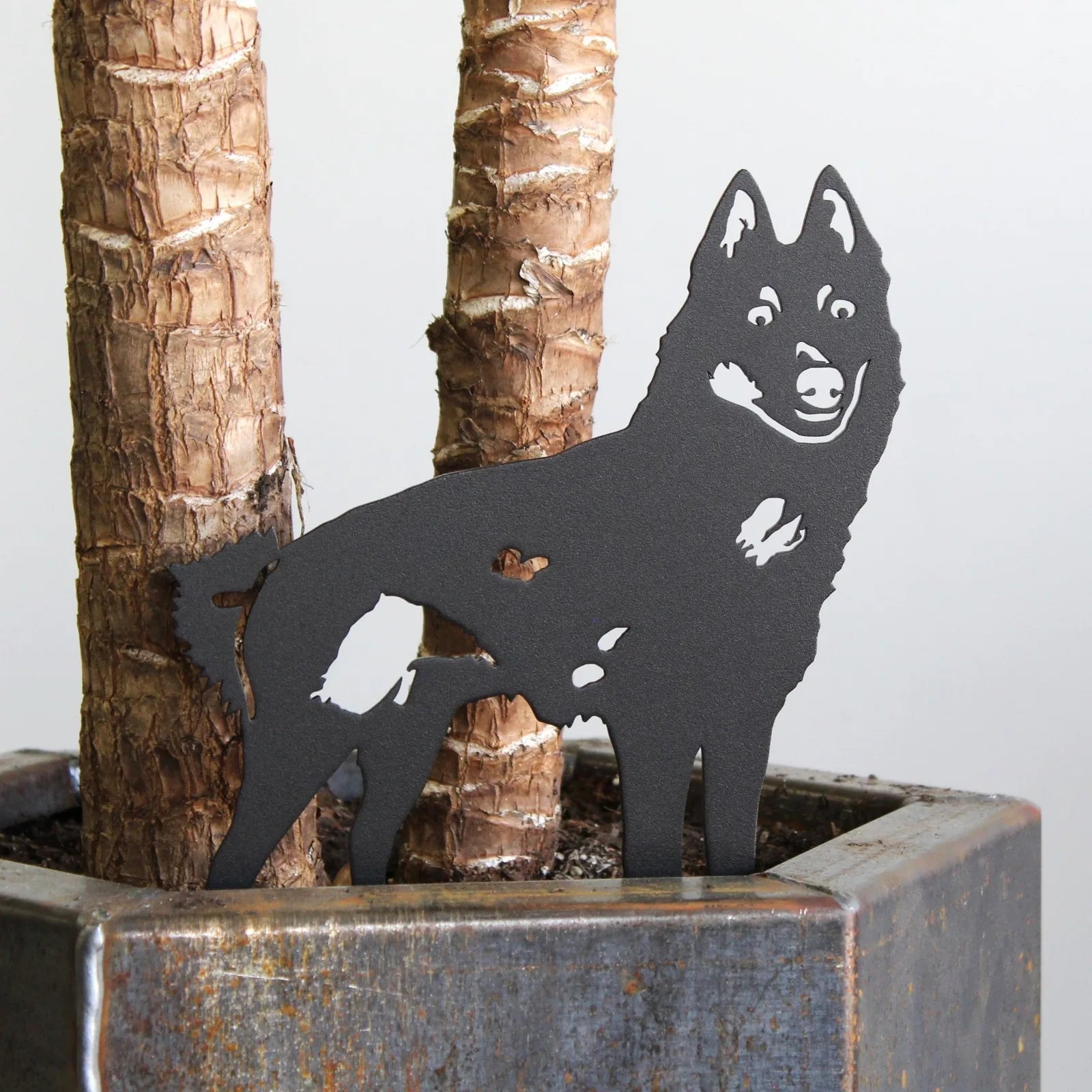 Black metal silhouette of a Schipperke dog, displayed next to a textured plant pot with tree trunks in the background.