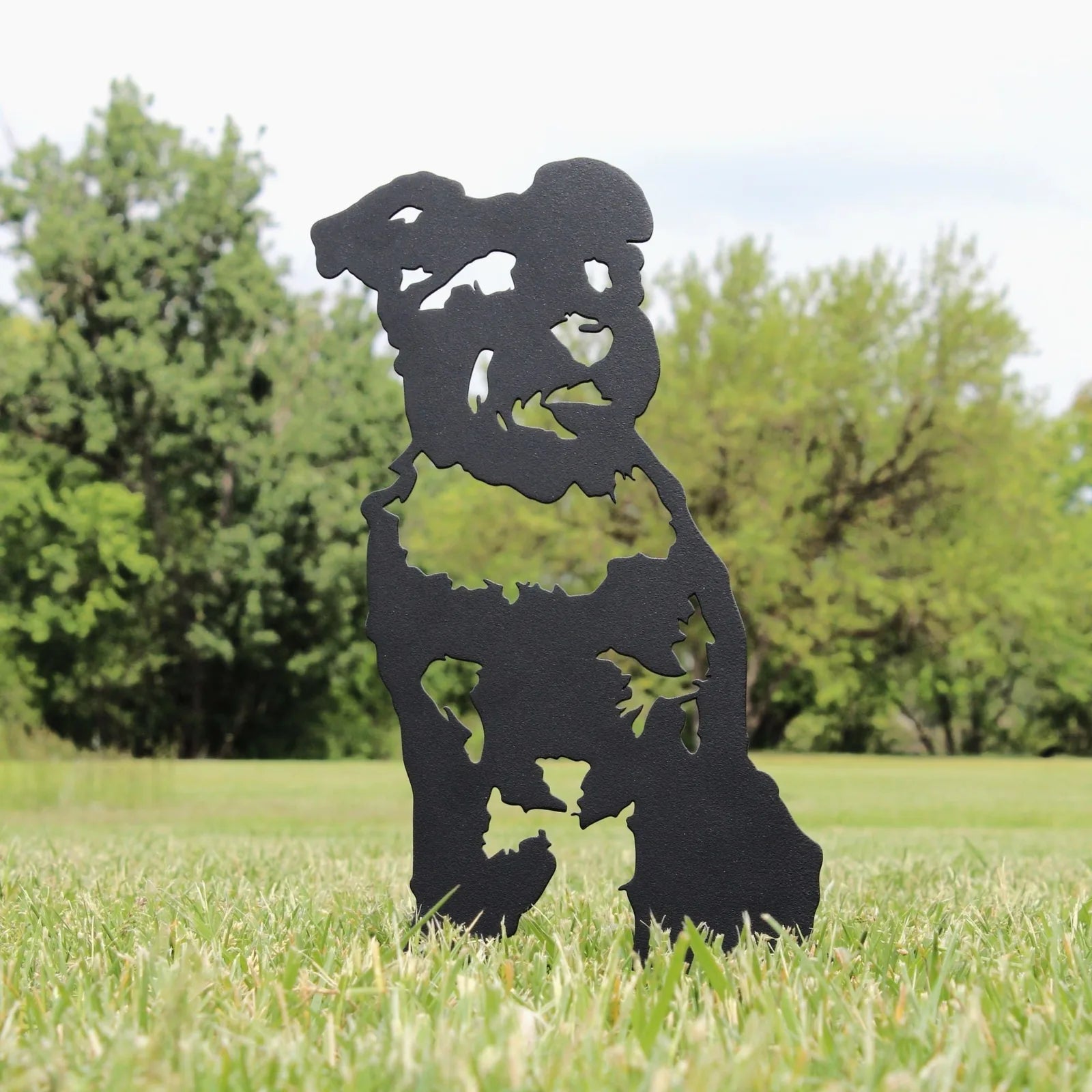 Schnauzer silhouette in matte black metal, standing in a grassy outdoor space with greenery in the background.