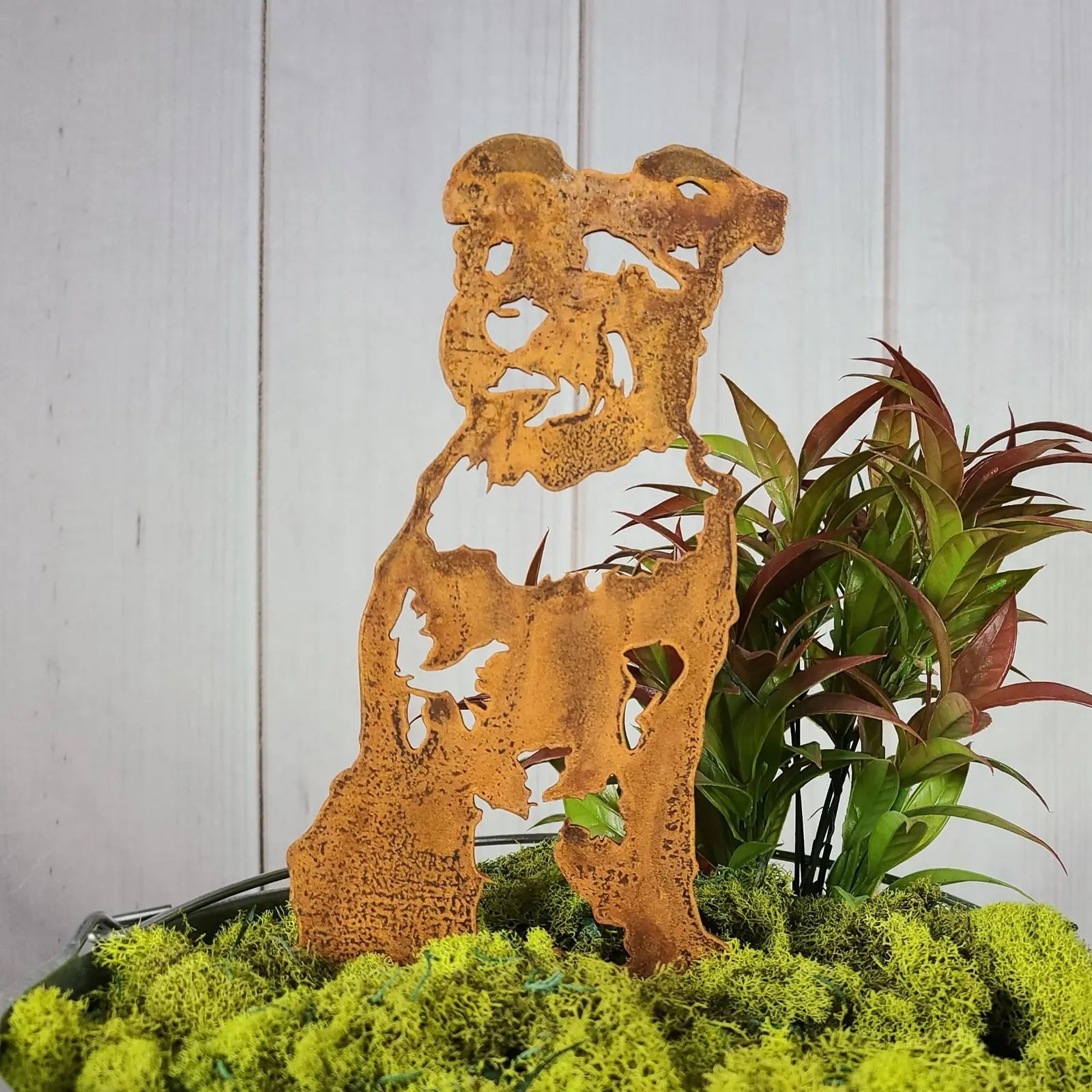Schnauzer metal silhouette in rust finish, set against green moss and foliage, perfect for outdoor decor.
