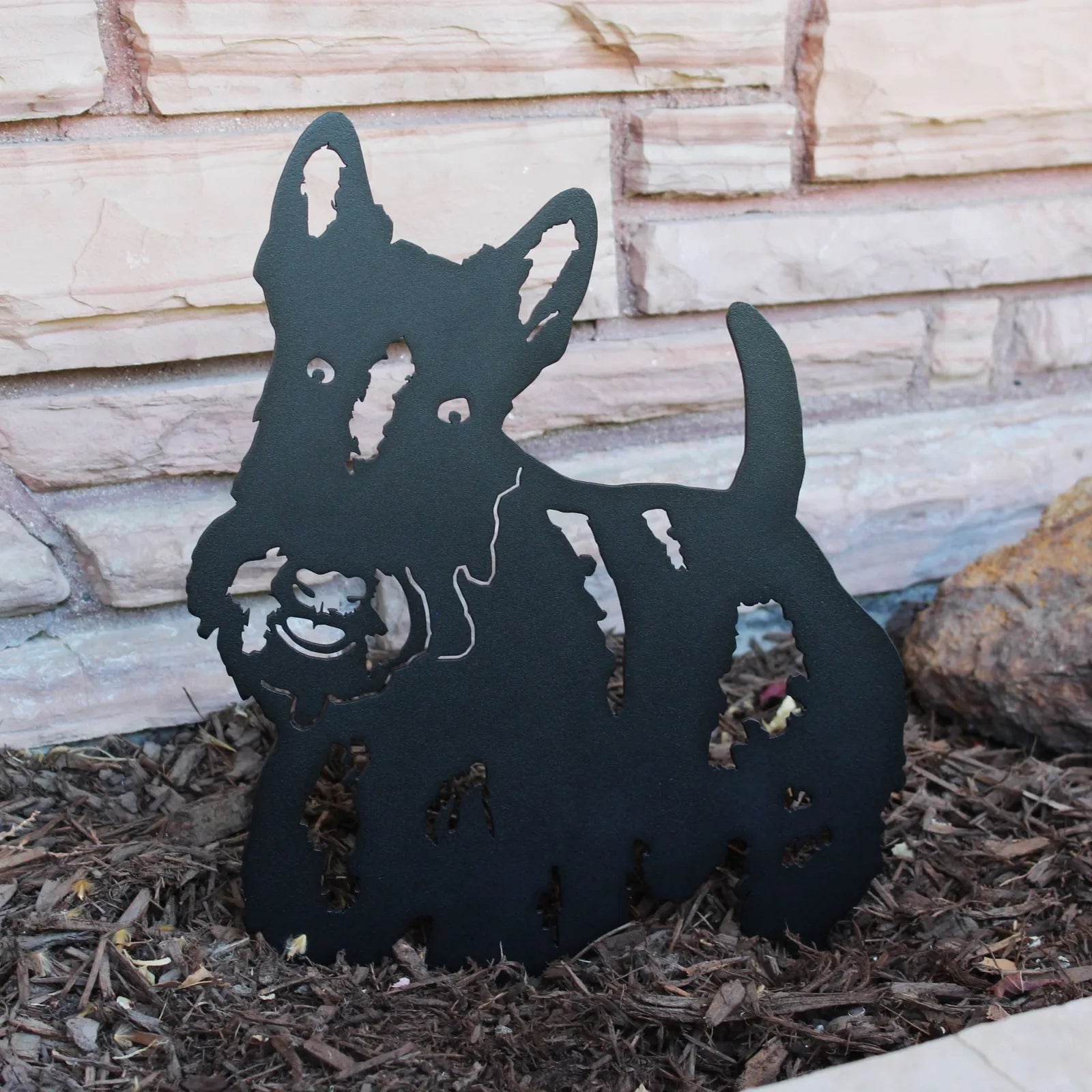 Black metal silhouette of a Scottish Terrier, featuring intricate cutouts, displayed in garden mulch against a stone wall.