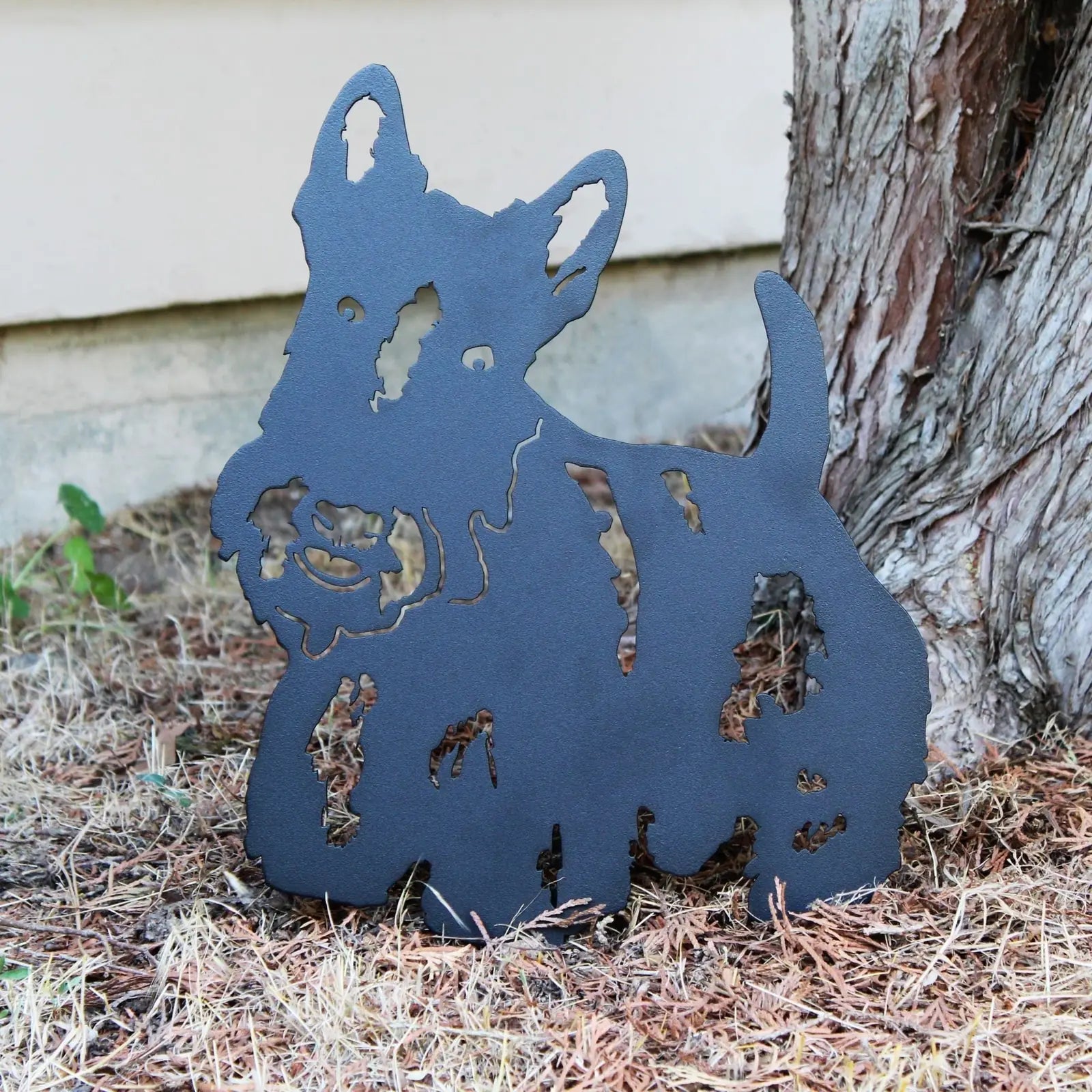 Black Scottish Terrier metal silhouette resting on dry ground, showcasing intricate cutout design for garden decor.