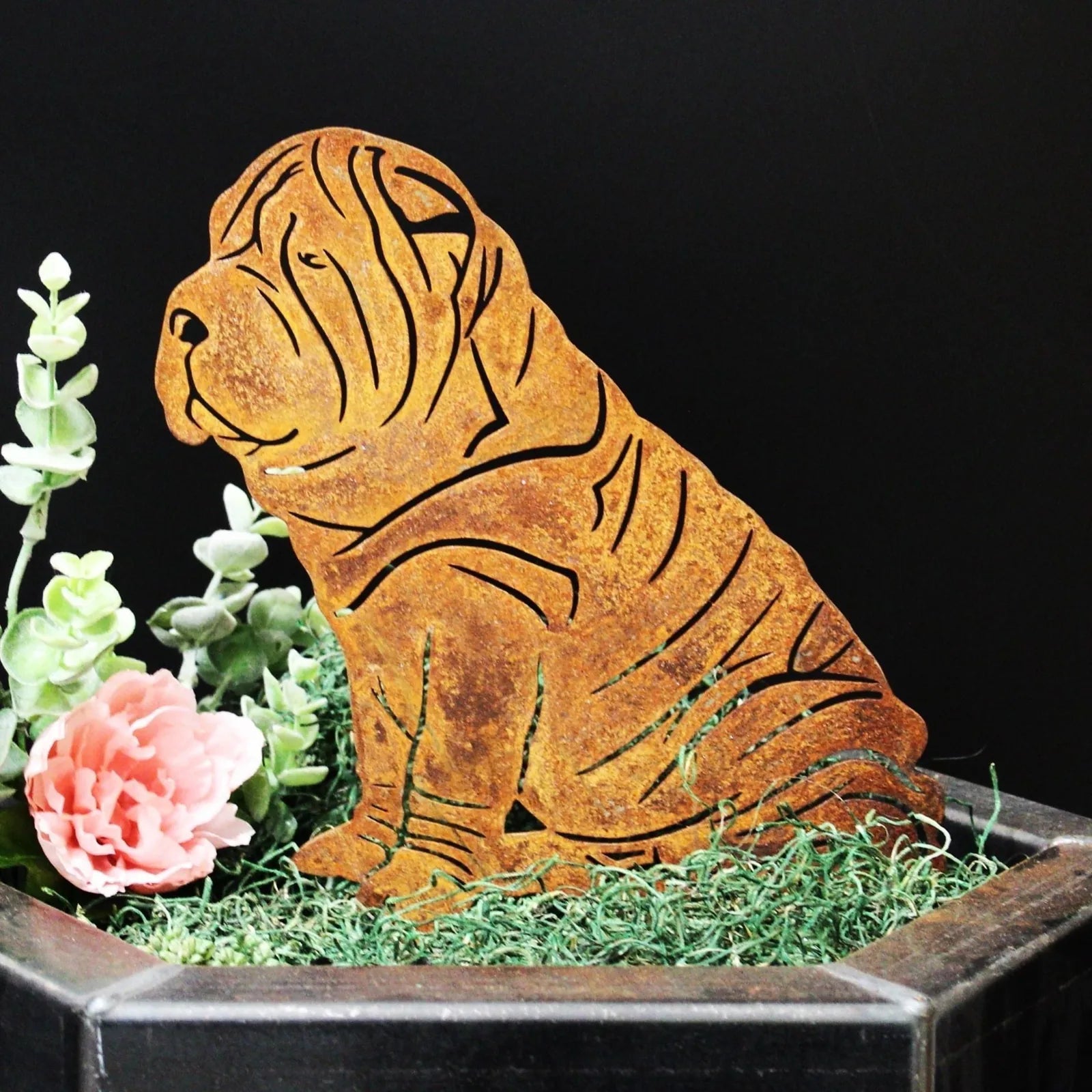 Rusty Corten steel silhouette of a Shar Pei dog, nestled among green grass and pink flowers in a planter.