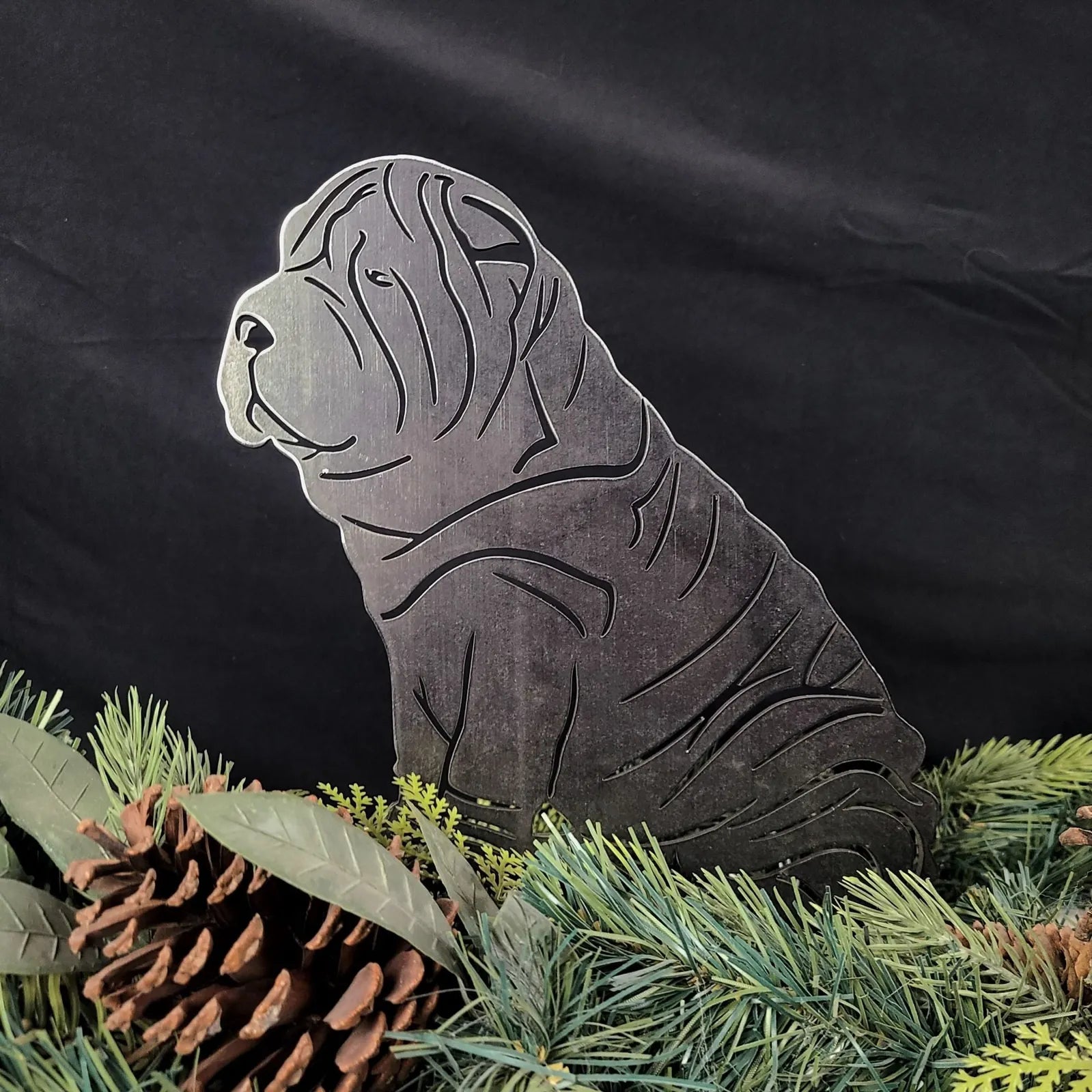 Shar Pei metal silhouette in matte black, nestled among greenery and pinecones, showcasing its intricate design.