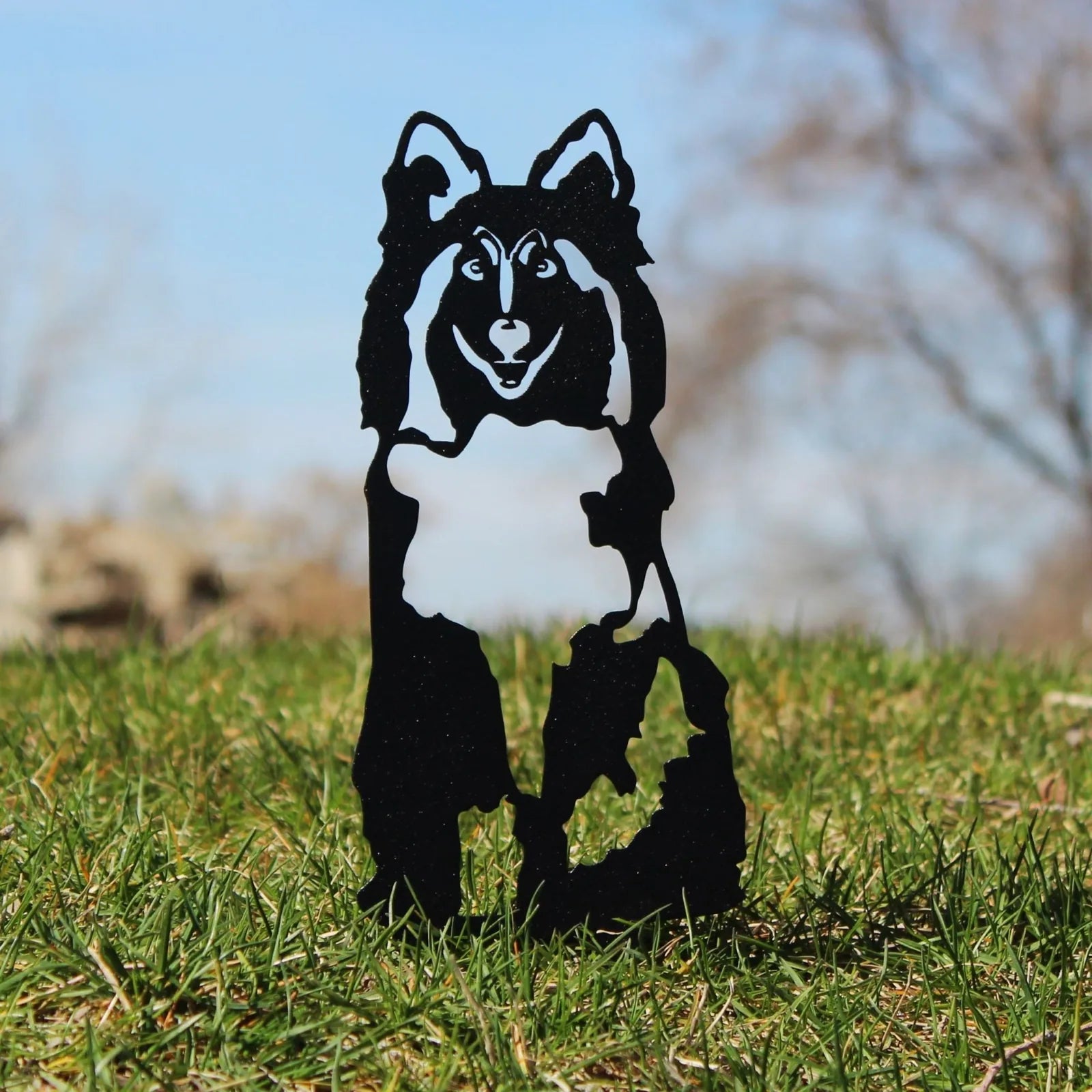 Black metal silhouette of a Shetland Sheepdog standing on grass with a blue sky background, showcasing intricate details.