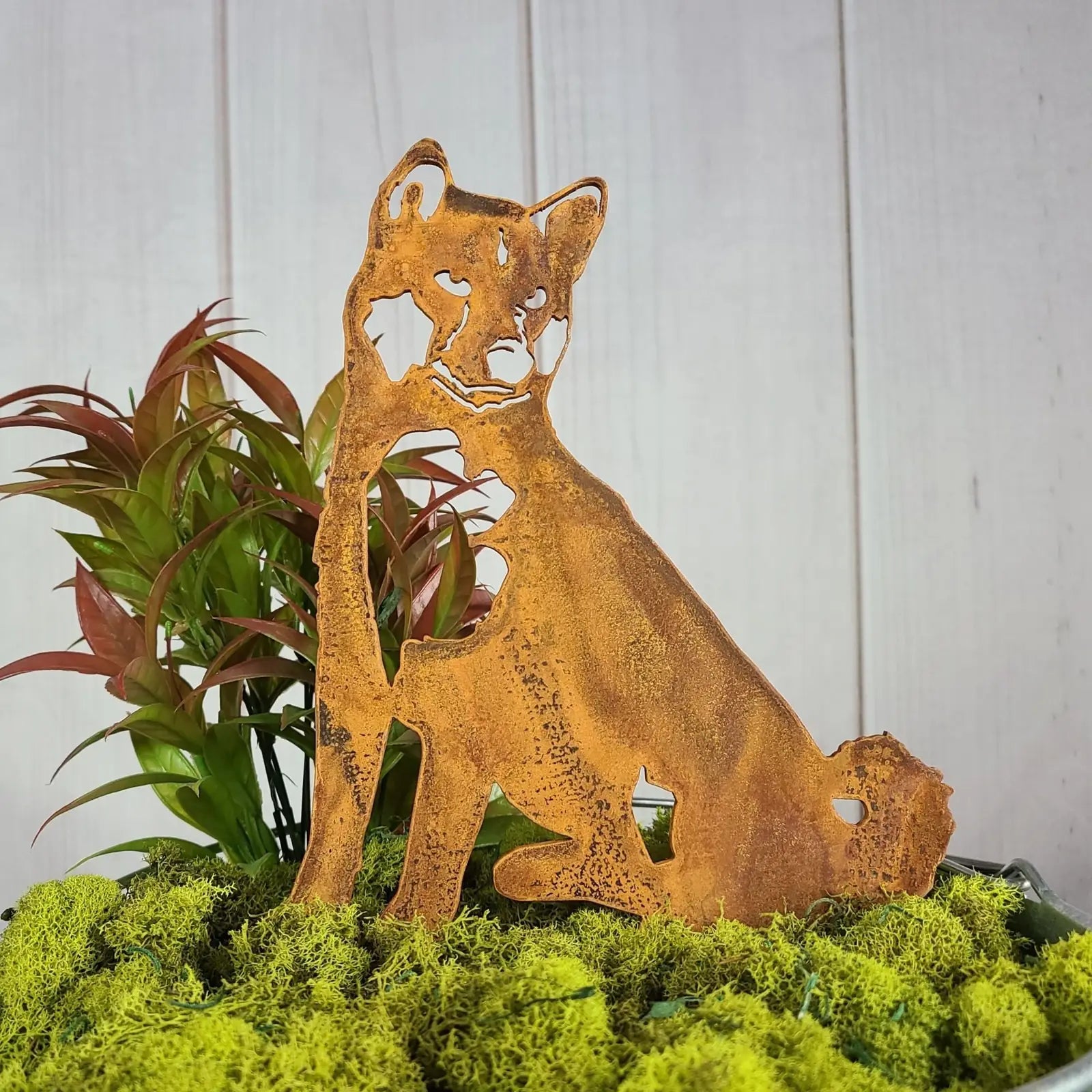 Shiba Inu metal silhouette in rusted Corten steel among vibrant green moss and foliage, ideal for outdoor decor.