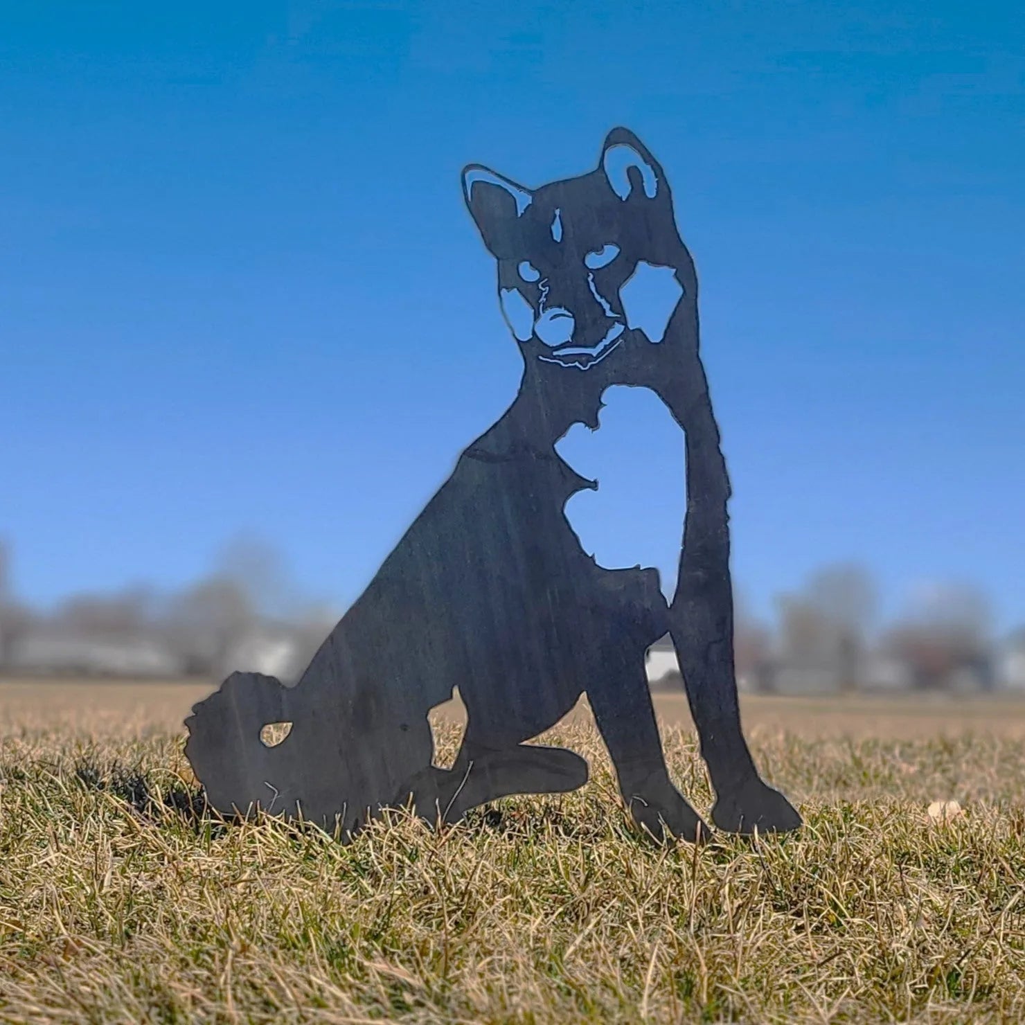 Handmade black metal silhouette of a Shiba Inu seated in grass against a clear blue sky. Perfect for outdoor decor.