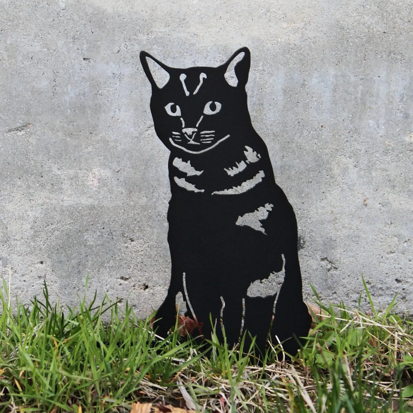 Black metal silhouette of a shorthair tabby cat amid grass and concrete, showcasing rustic outdoor decor.
