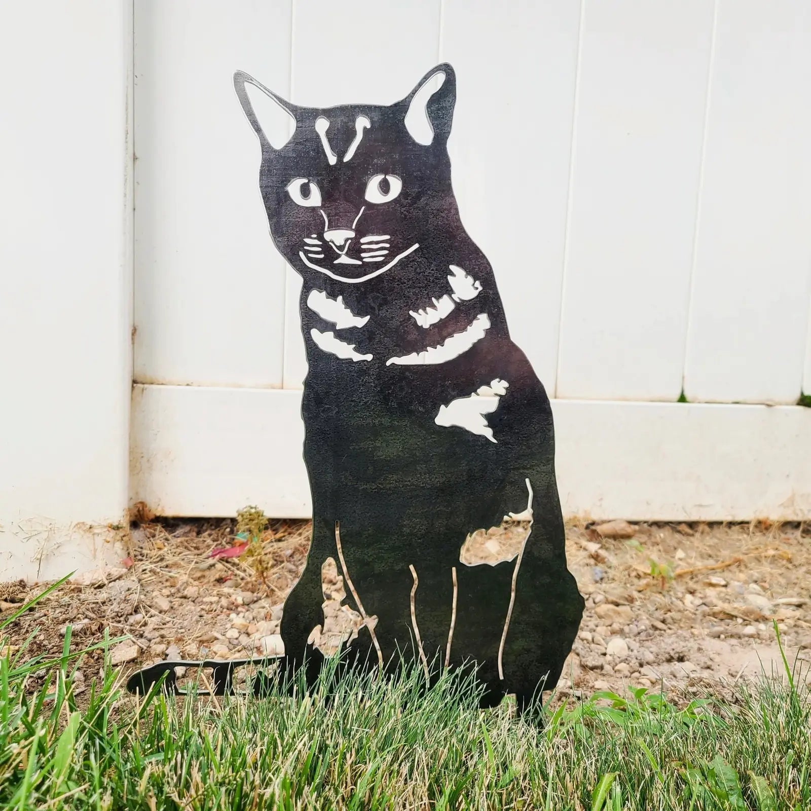 Black metal silhouette of a shorthair tabby cat, showing distinct features, placed in grass near a white fence.