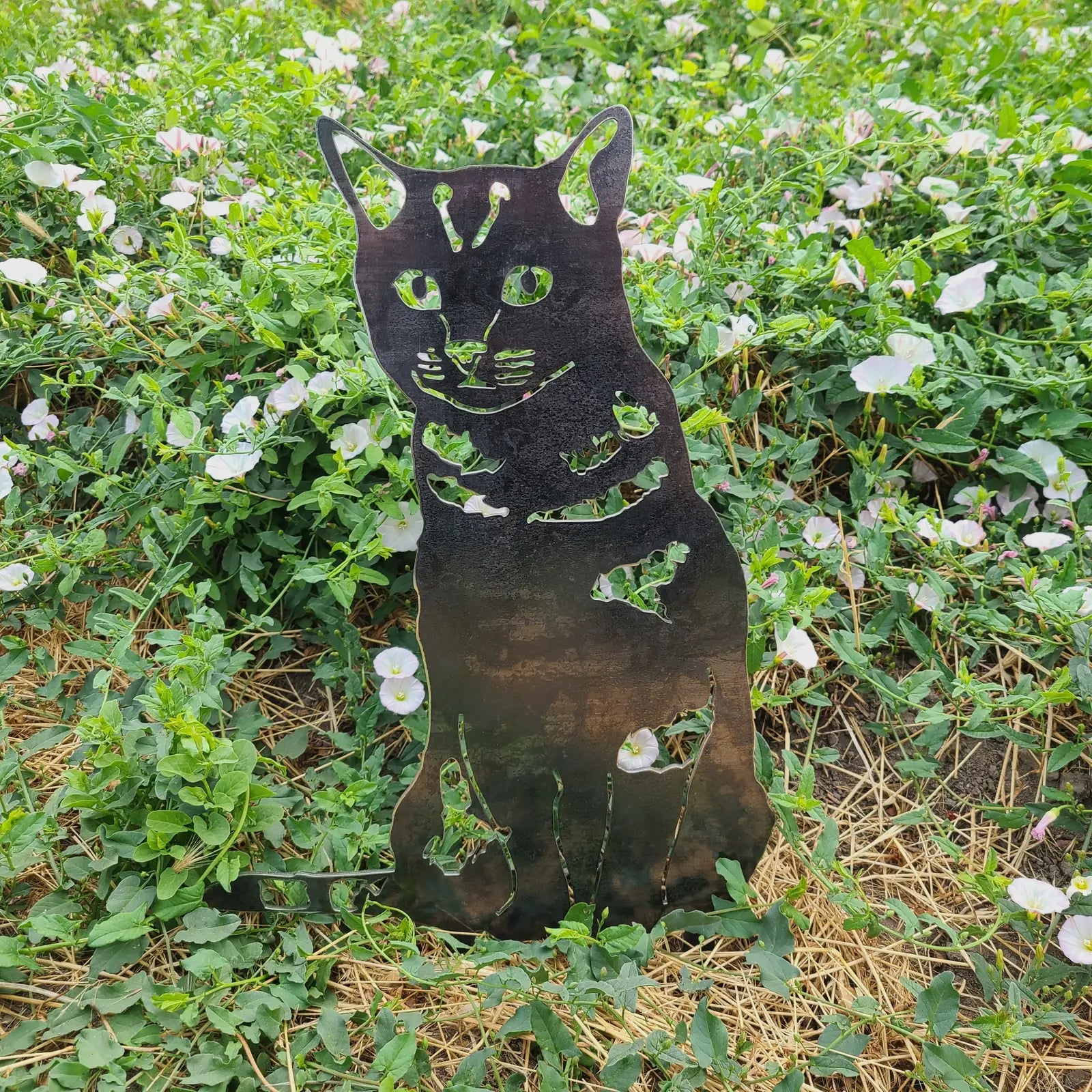 Shorthair tabby cat silhouette in black metal, surrounded by green foliage and white flowers, enhancing outdoor decor.