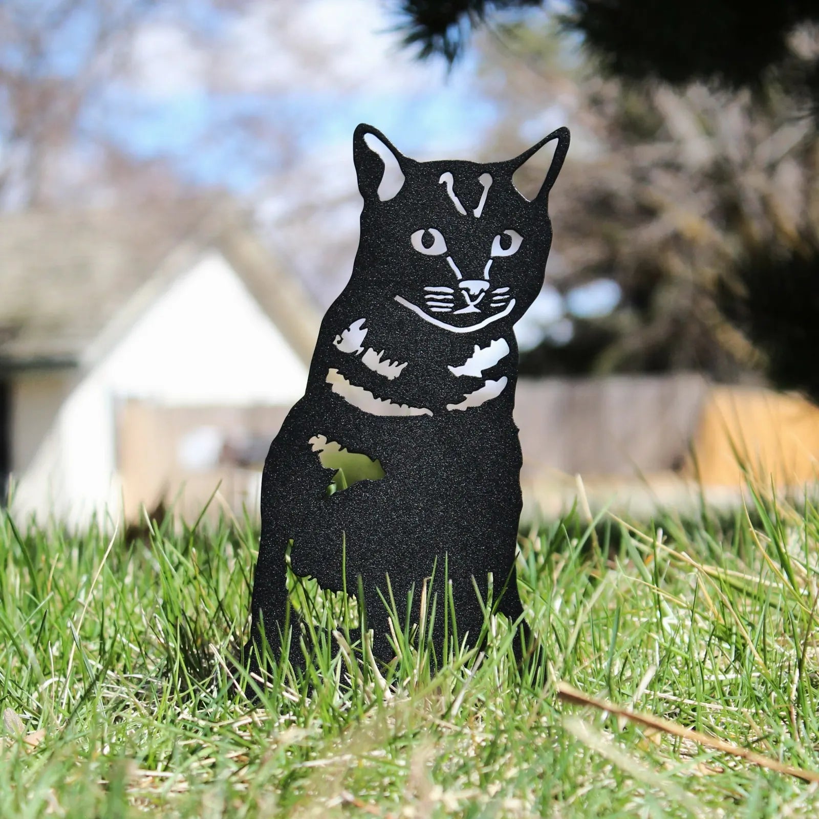 Black metal silhouette of a shorthair tabby cat, perched on grass in an outdoor setting, showcasing rustic design.
