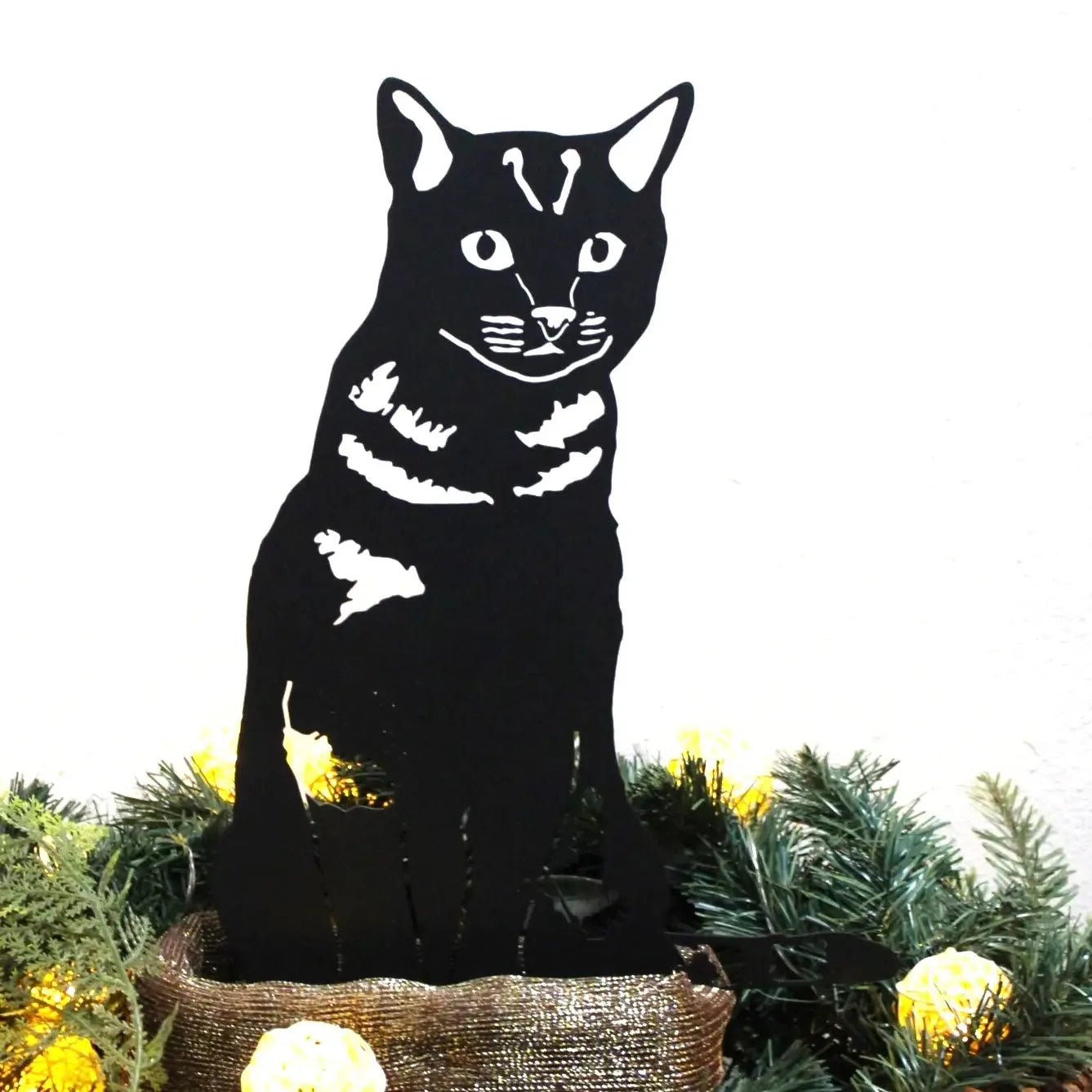 Black metal silhouette of a Shorthair Tabby cat, showcasing detailed facial features and stripes, set among greenery.