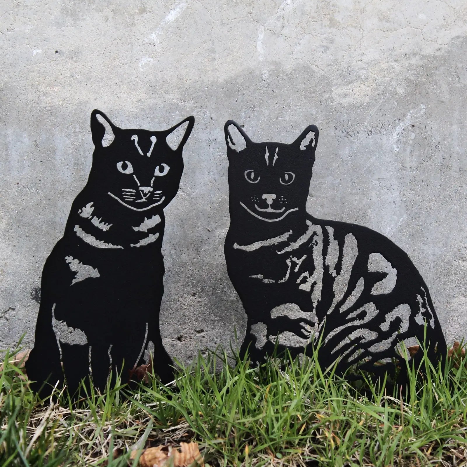 Black metal cat silhouettes, showcasing a shorthaired tabby design, placed against a concrete background with grass.