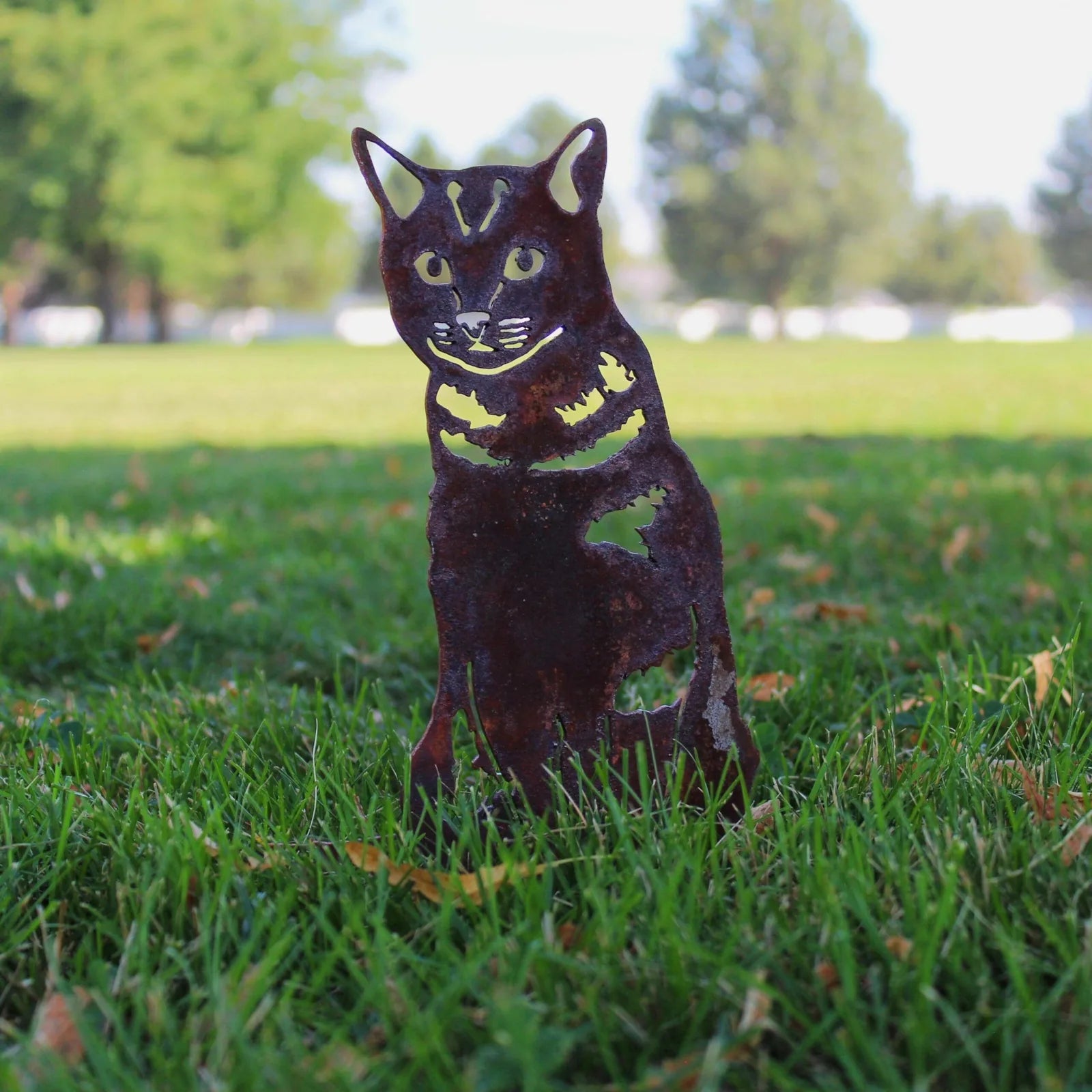 Rustic shorthair tabby cat silhouette in weathered steel, set against a grassy outdoor backdrop.