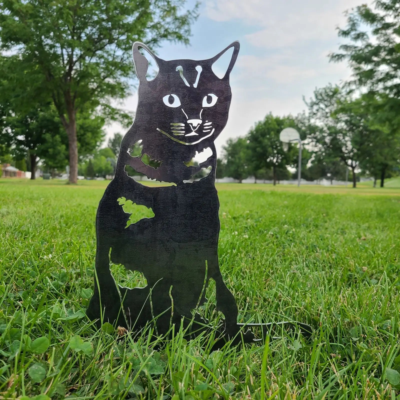 Black metal silhouette of a shorthair tabby cat set on green grass in a park, showcasing its detailed design.