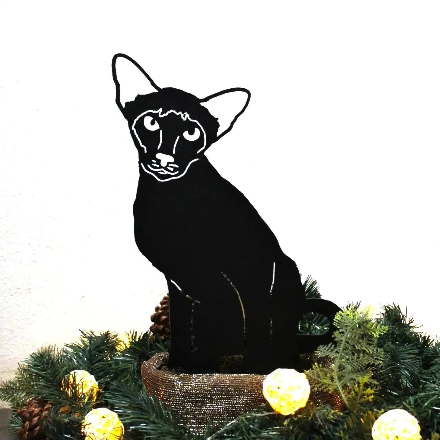 Black Siamese cat silhouette in metal, featuring detailed facial lines, displayed amidst festive greenery and lights.