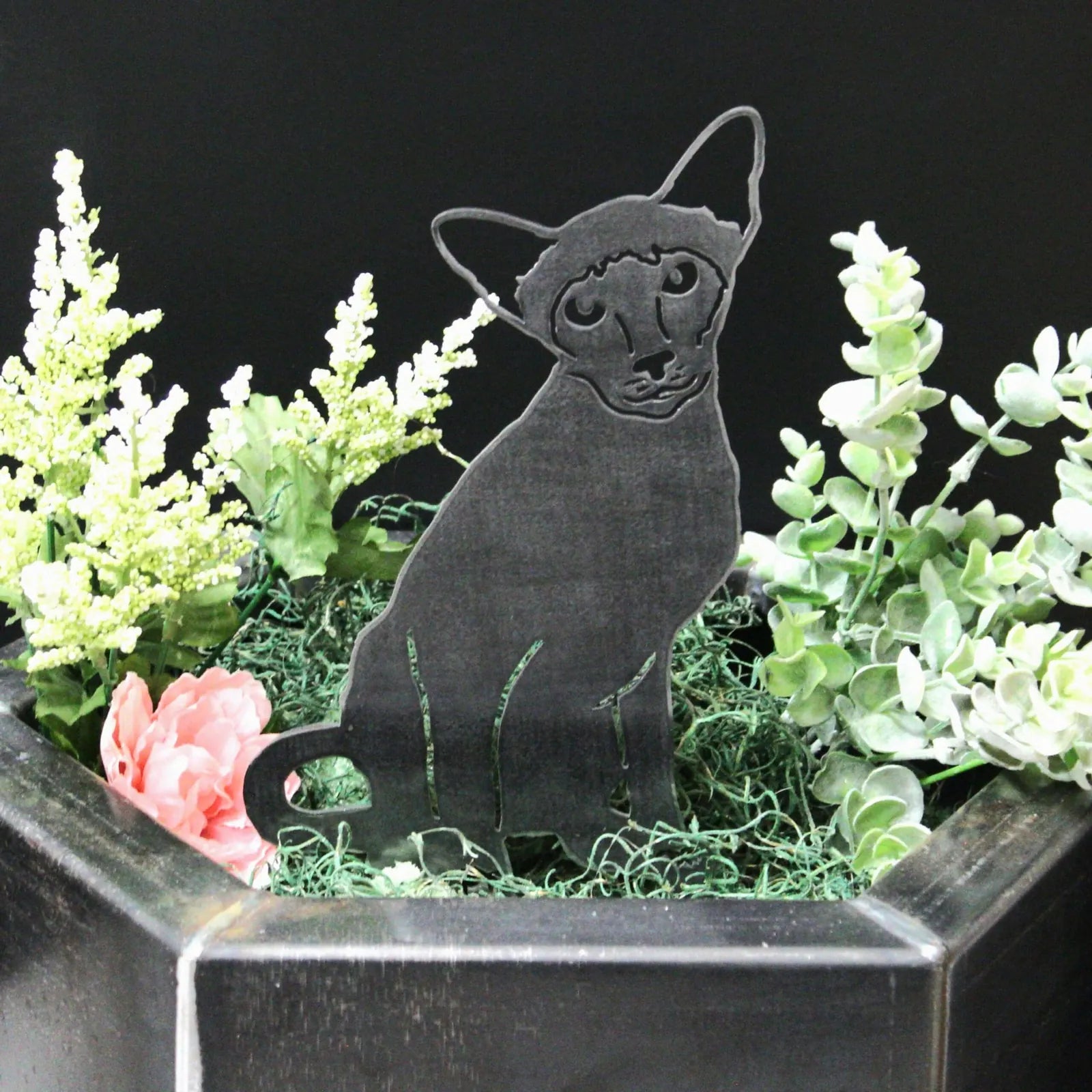 Handmade black metal Siamese cat silhouette nestled among green foliage and pink flowers in a decorative planter.