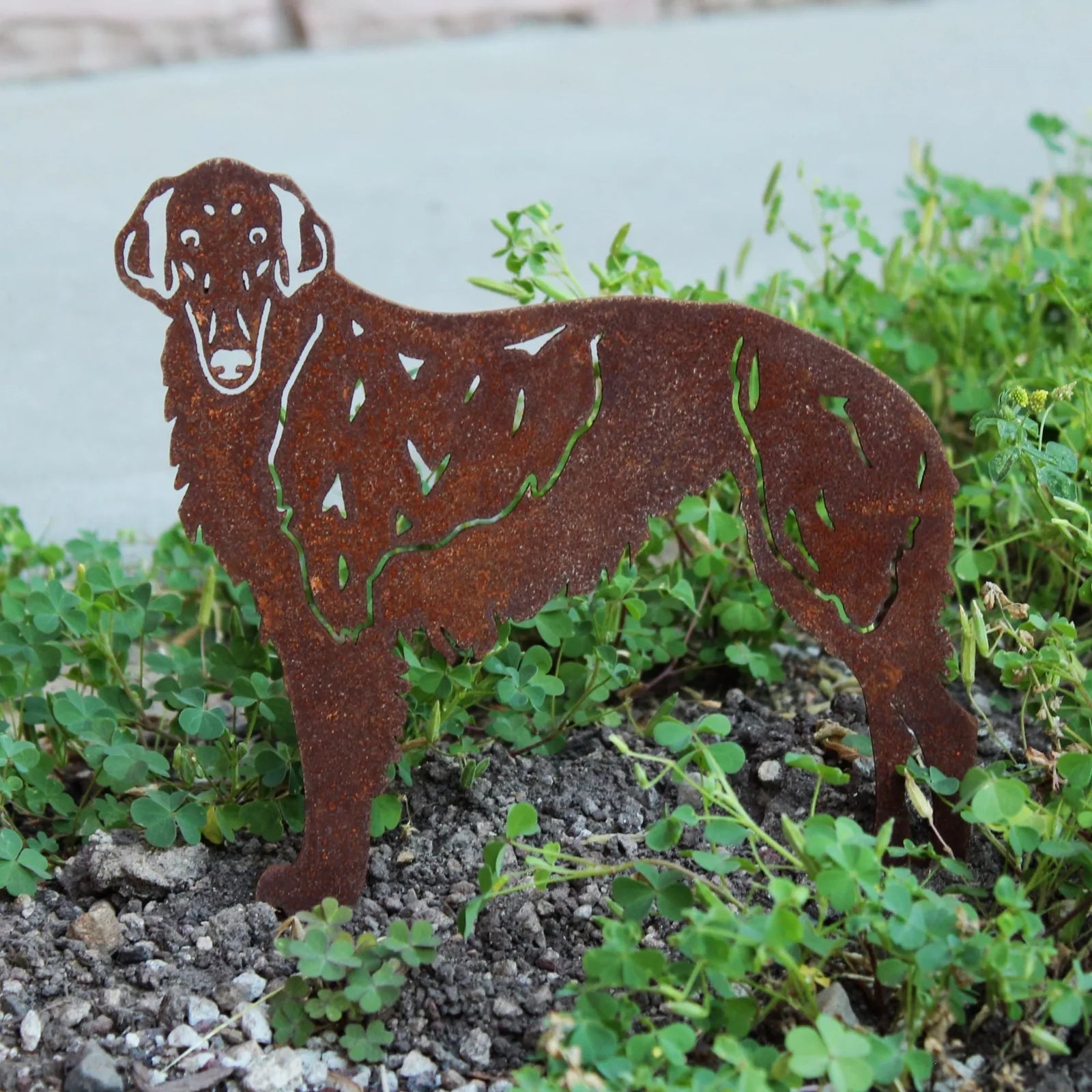 Rust-colored Silken Windhound metal silhouette art stands among green plants in a decorative outdoor setting.
