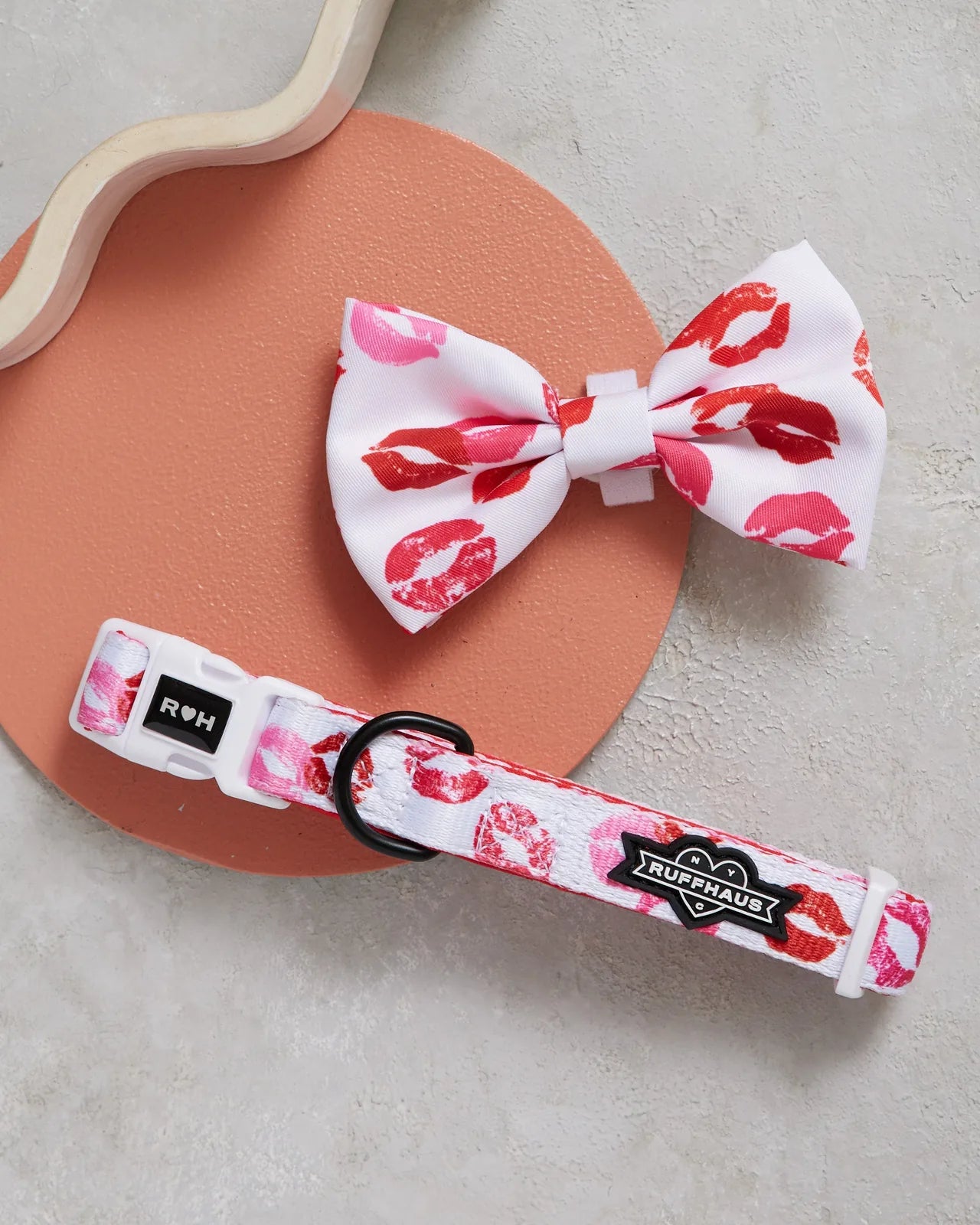 White dog collar with pink and red kiss print alongside a matching bowtie, resting on a coral background.