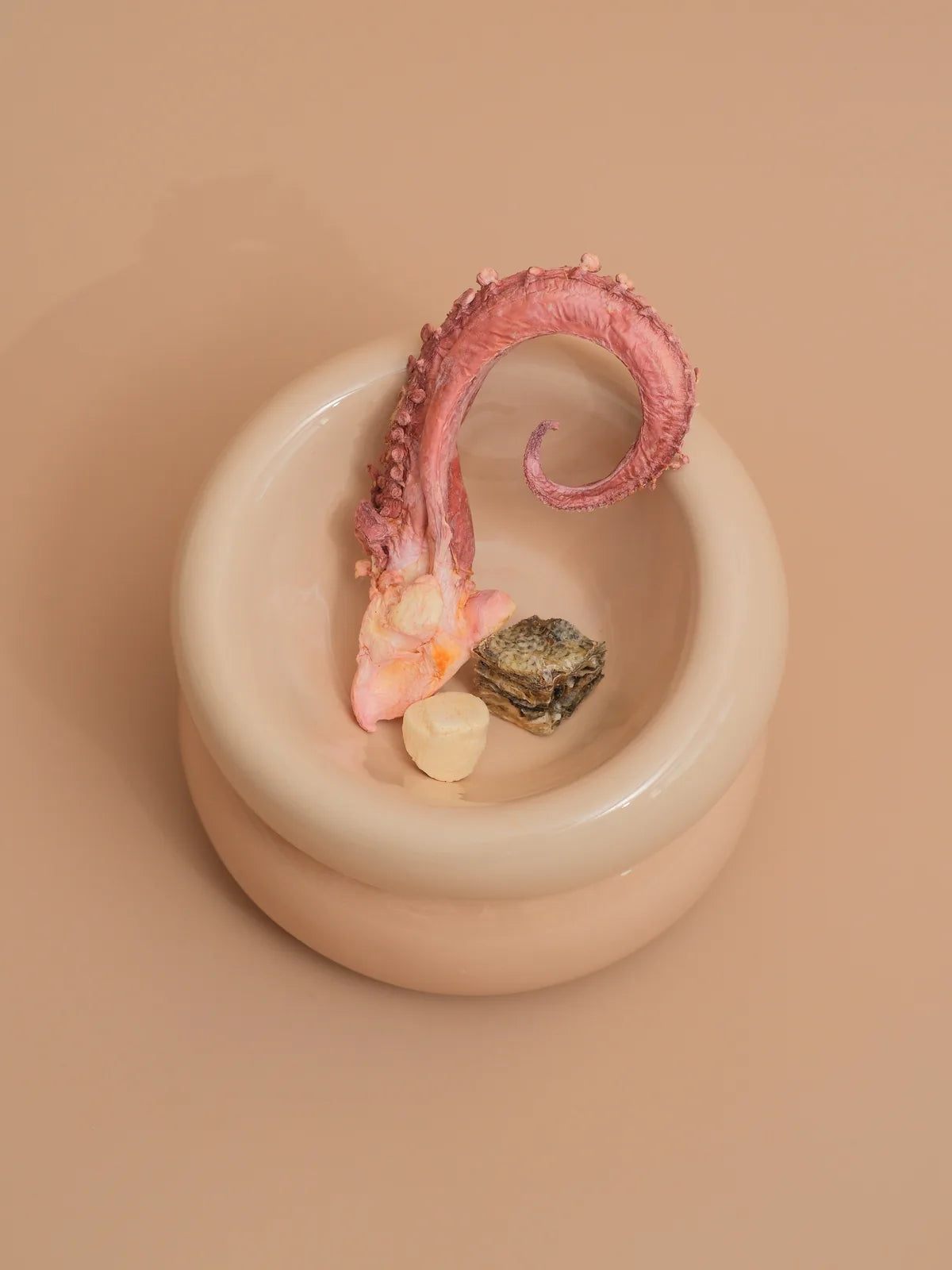 Cream beige pet bowl with a diagonal opening, featuring an octopus design and assorted colorful treats inside.