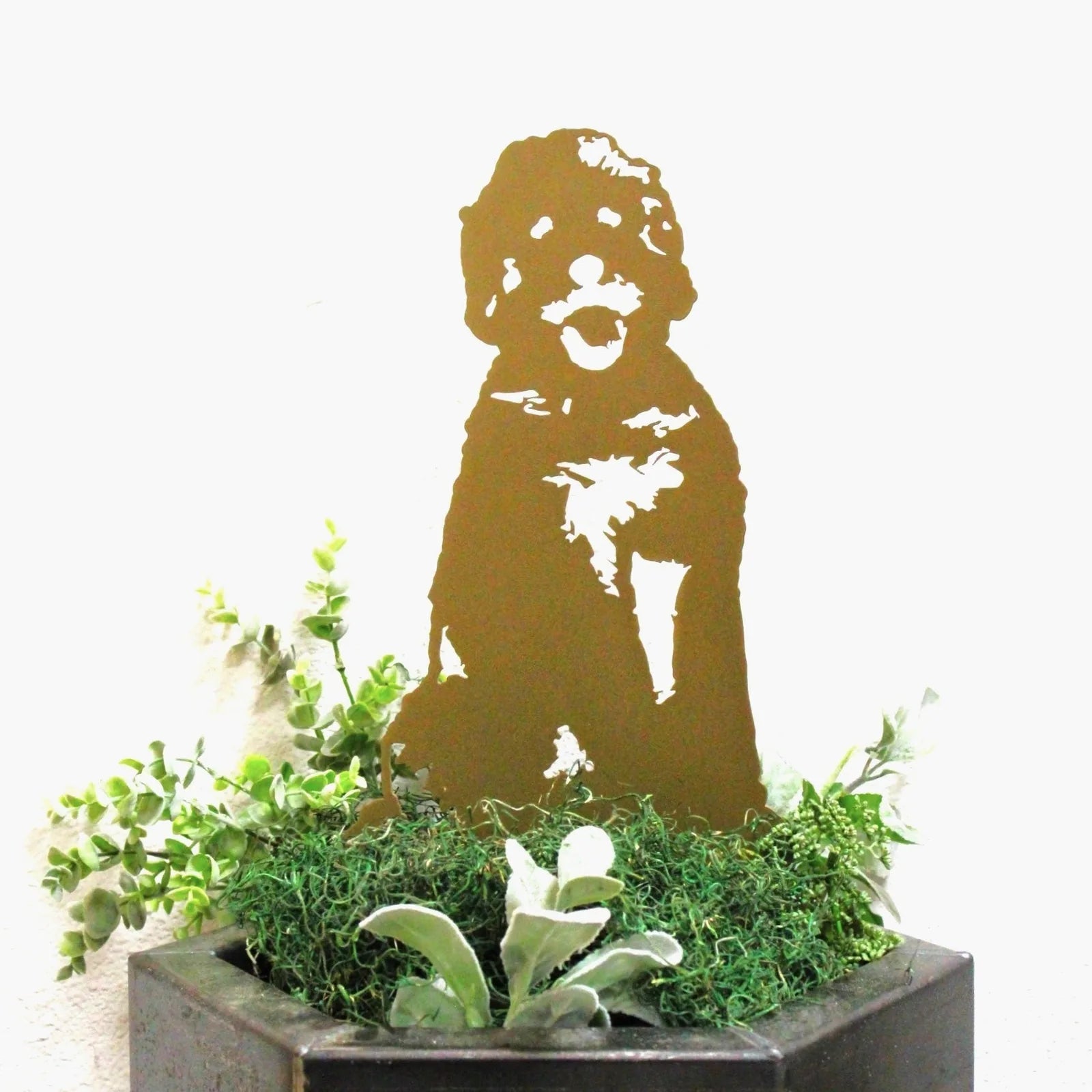Gold Doodle metal silhouette art piece in a subtle metallic finish, surrounded by green foliage in a planter.