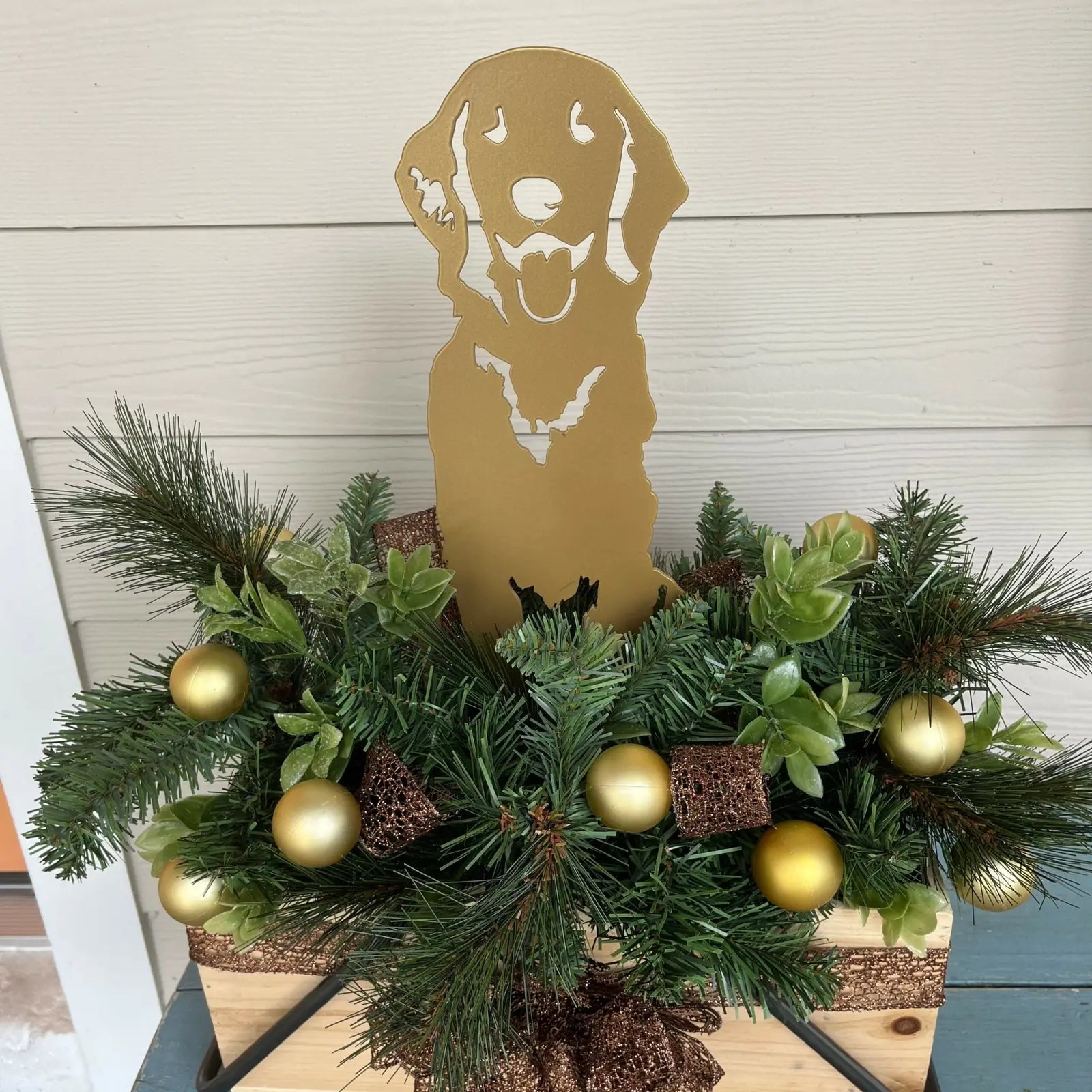 Golden retriever silhouette in gold metal, surrounded by greenery and decorative golden accents in a festive arrangement.