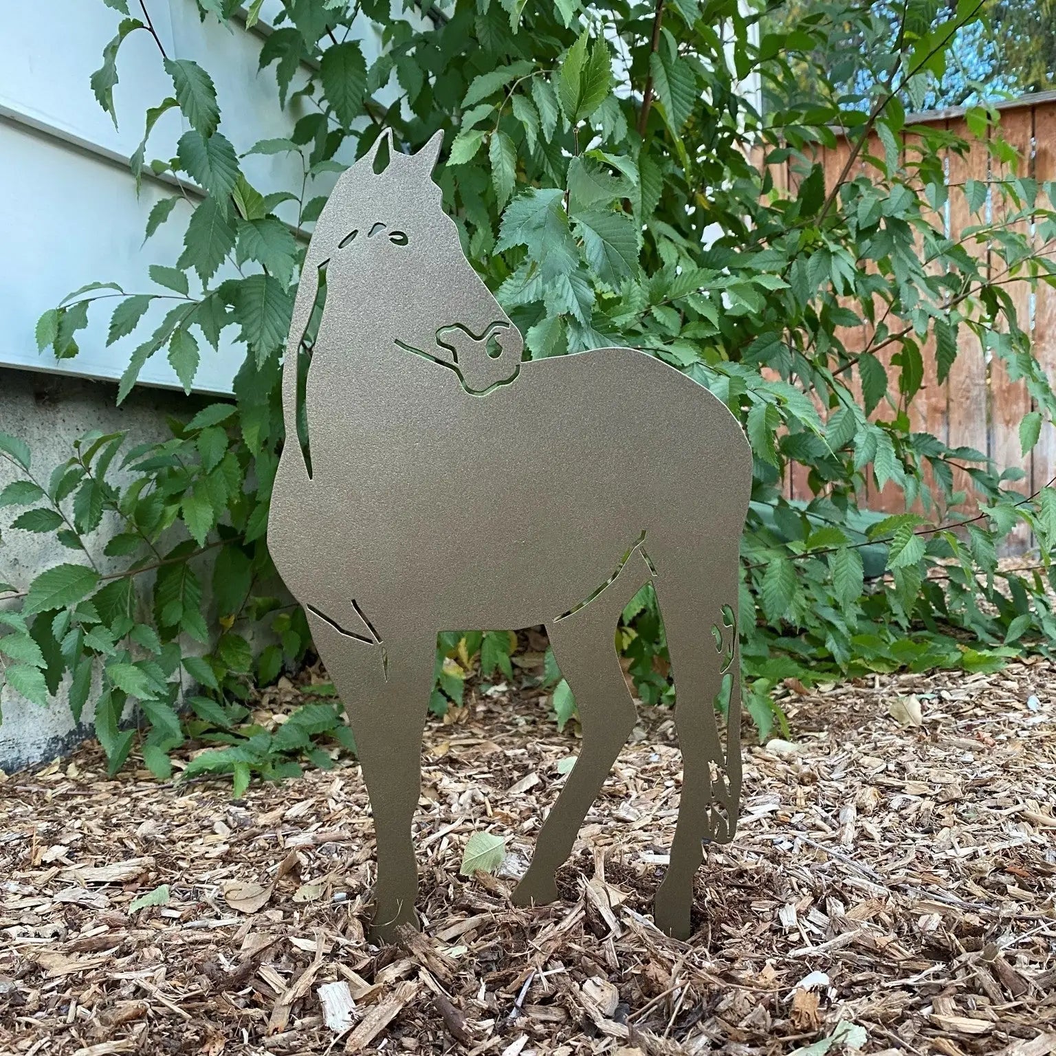 Gold metal horse silhouette standing among greenery on mulch, showcasing stable, artistic design for outdoor decor.