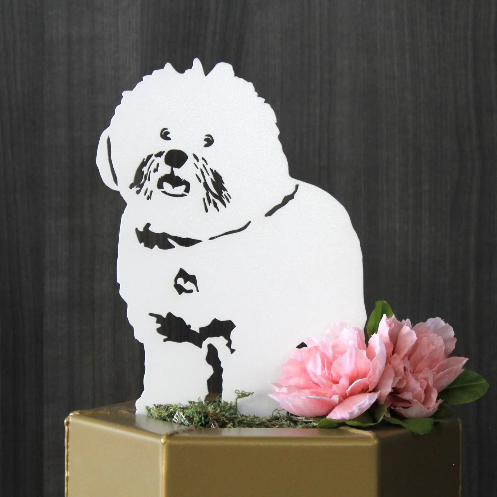 White Bichon Frise metal silhouette with flowers and moss on a gold surface against a dark wood background.