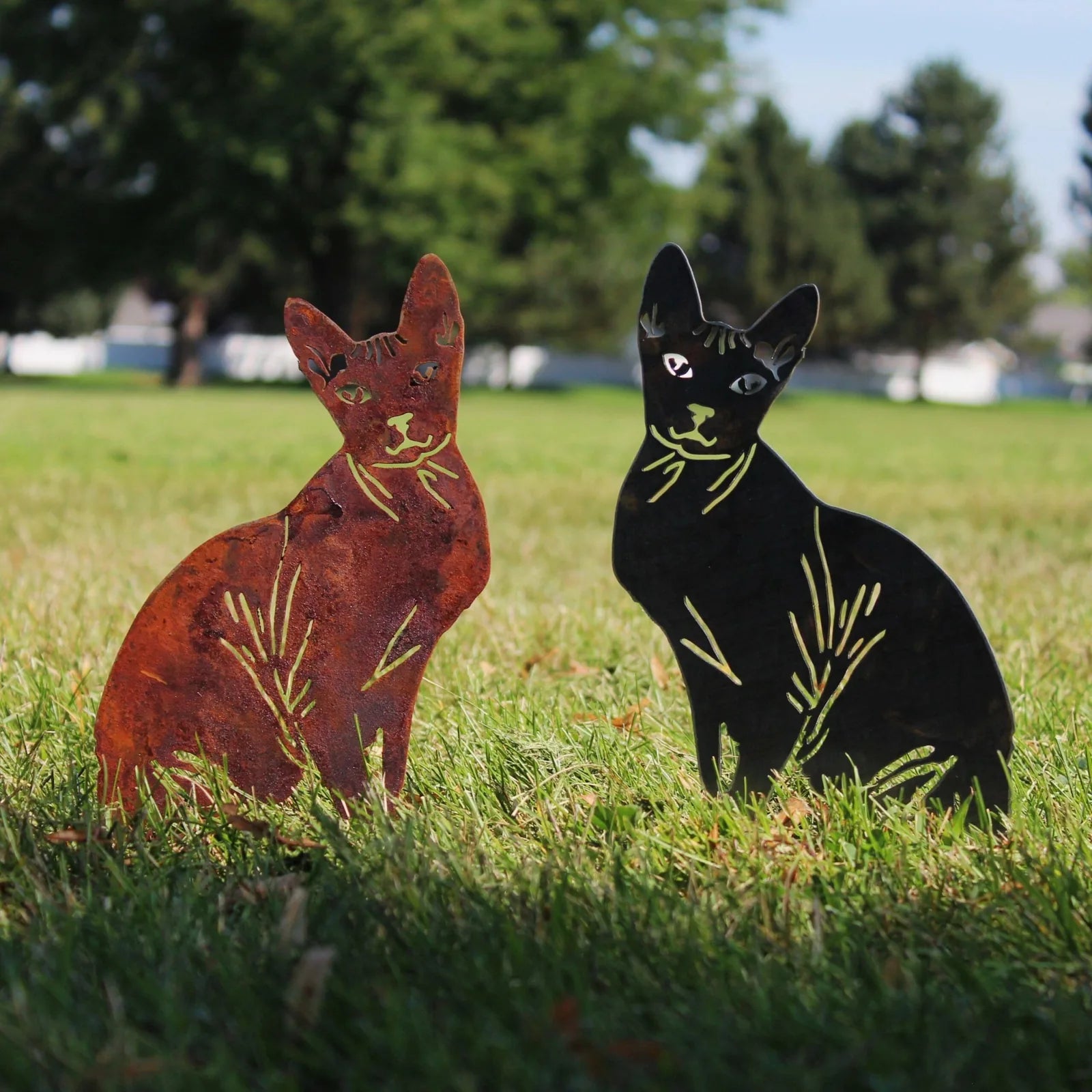 Rust-colored Corten steel and black powder-coated metal Sphynx cat silhouettes standing on grass in a garden setting.