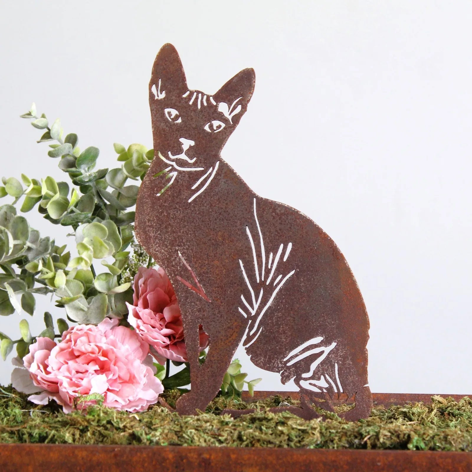 Sphynx cat silhouette made of weathered Corten steel, with pink flowers and greenery in a decorative arrangement.
