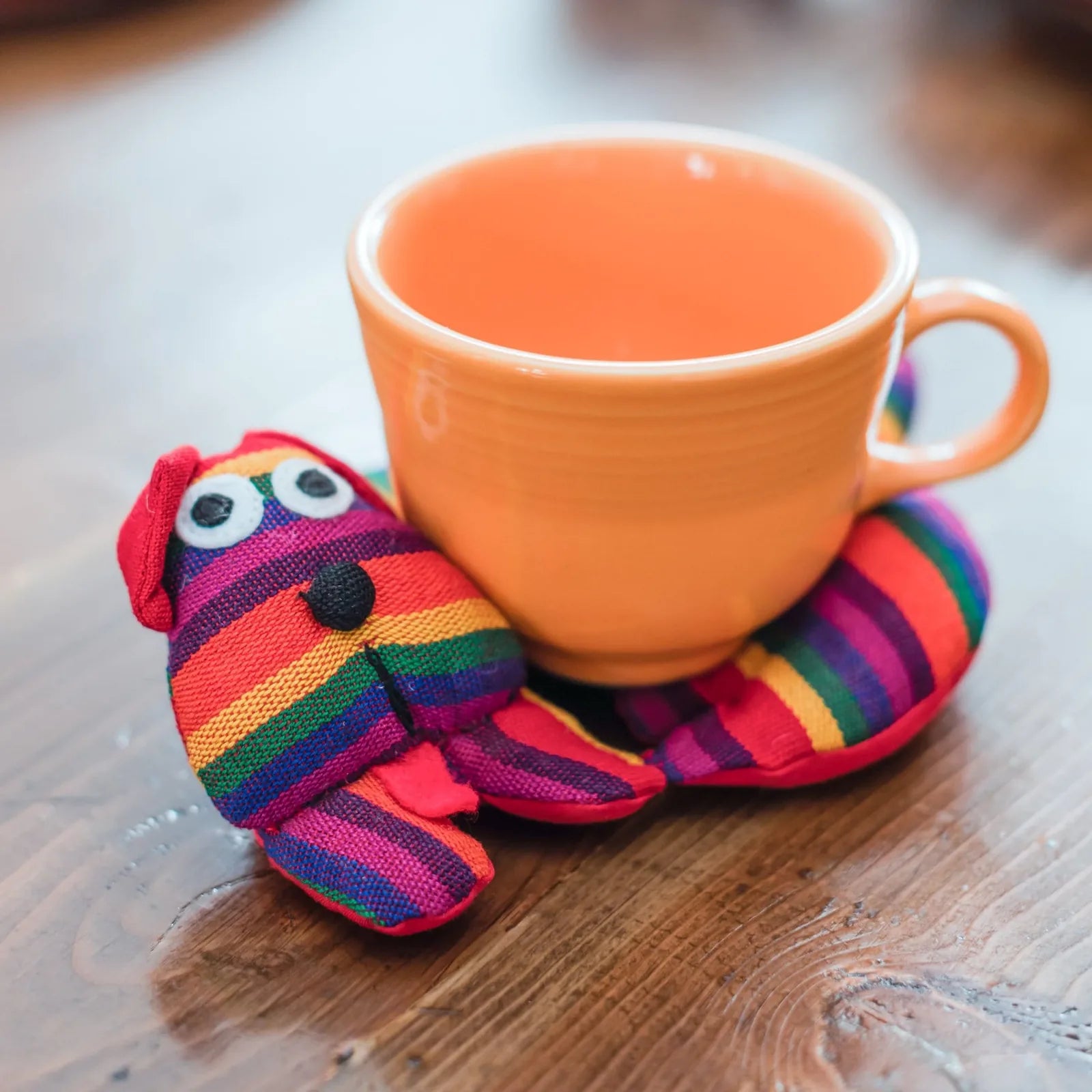 Colorful striped dog trivet in red, purple, orange, and green, cradling an orange cup on a wooden table.