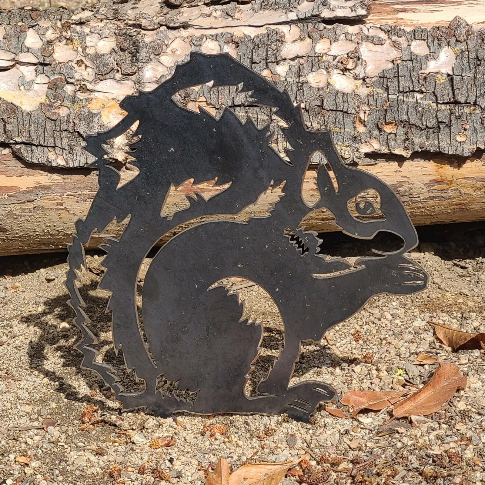 Black metal silhouette of a squirrel, featuring intricate cutouts, set against natural surroundings and textured wood.