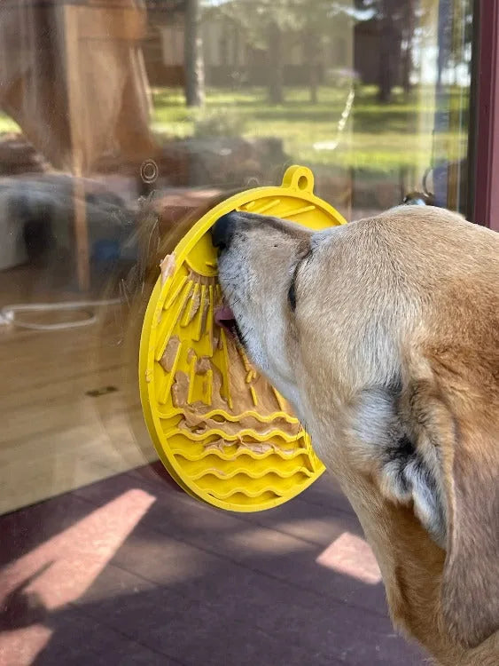 Labrador licking a yellow, beach-themed enrichment lick mat with textured surface, attached to a glass door.
