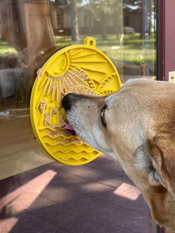 Dog licking peanut butter from a yellow beach-themed enrichment lick mat secured on glass, featuring sunny designs.